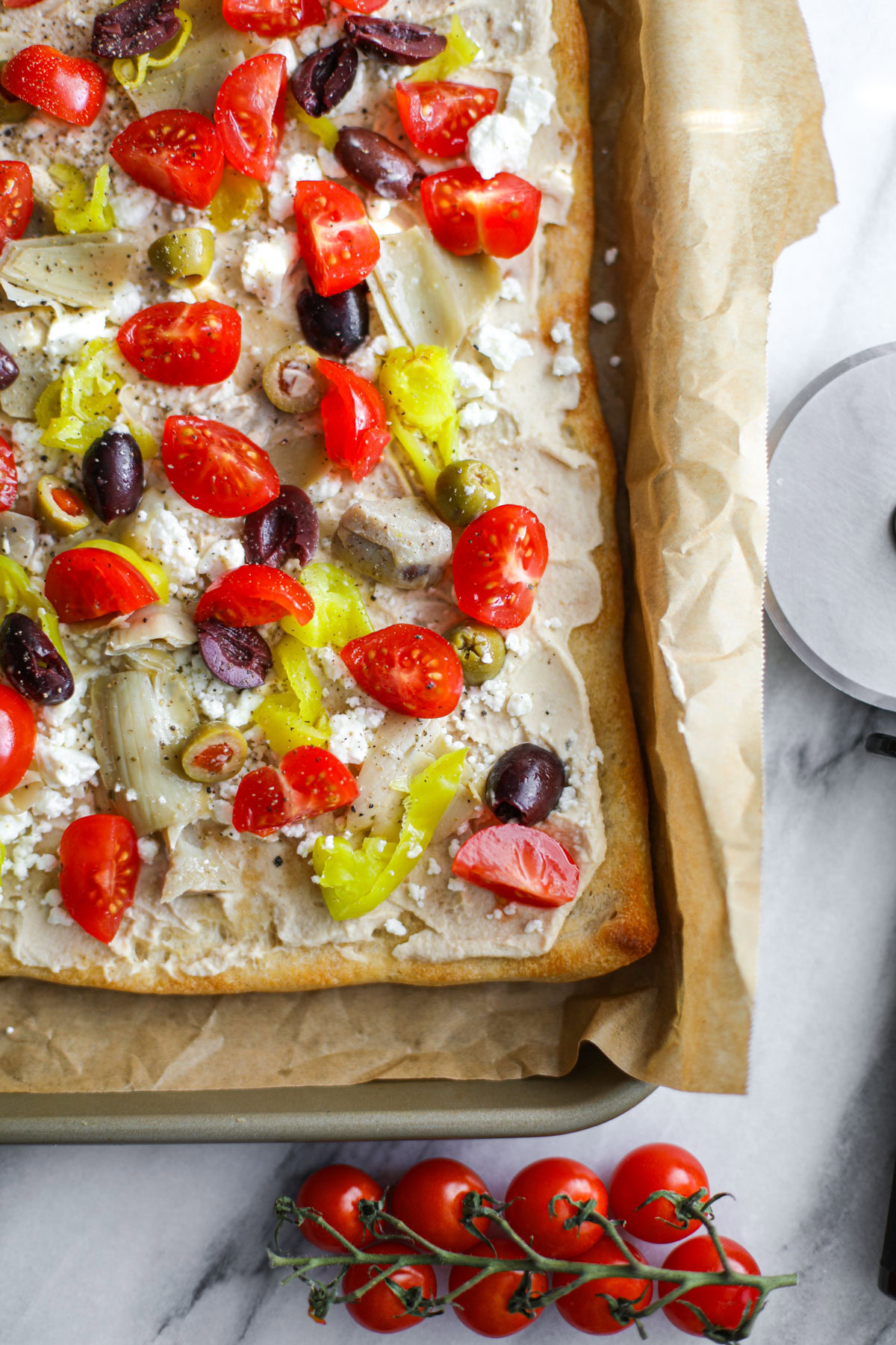A mediterranean pizza sitting on a marble cutting board topped with hummus, feta, cherry tomatoes, olives, artichoke hearts, and red onion with loose olives in the background.