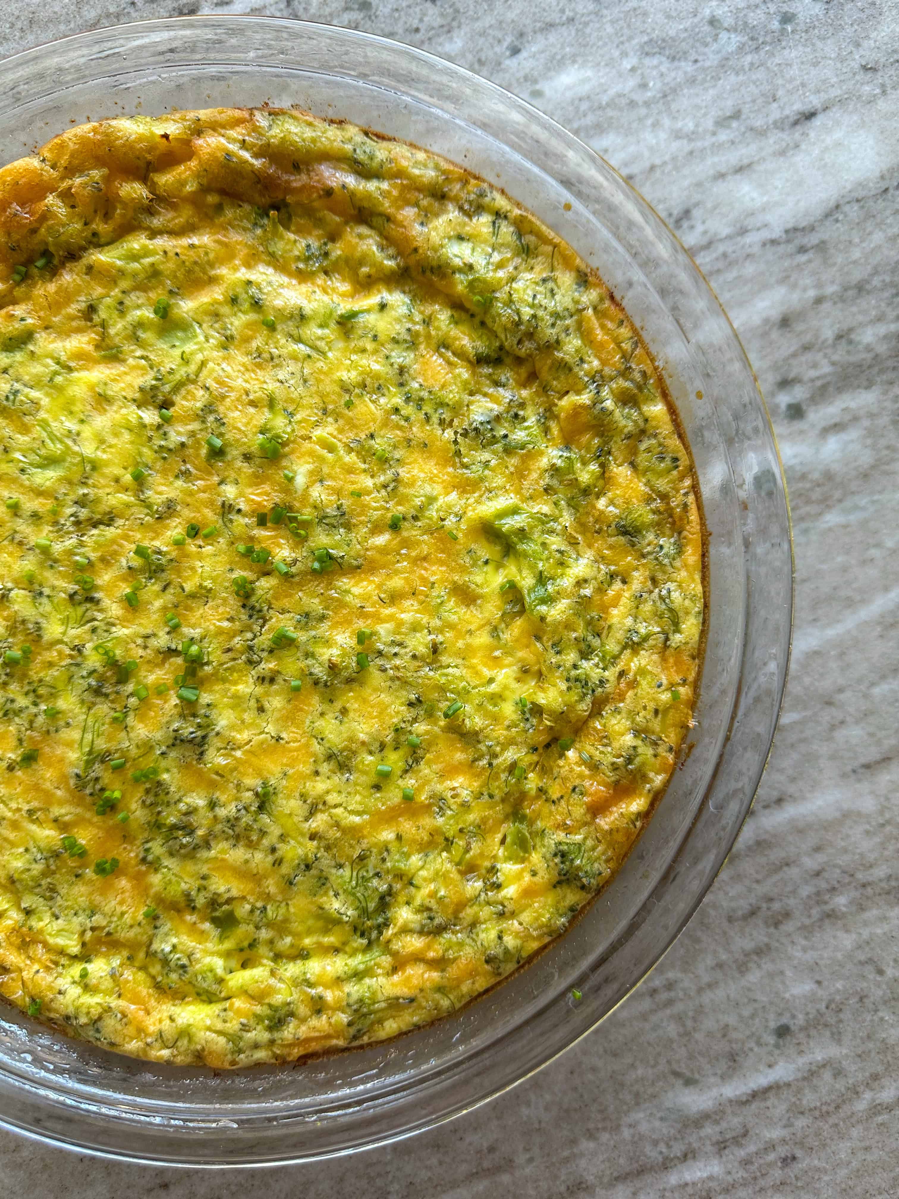 A broccoli and cheese quiche topped with chives in a glass pie dish on the countertop. 