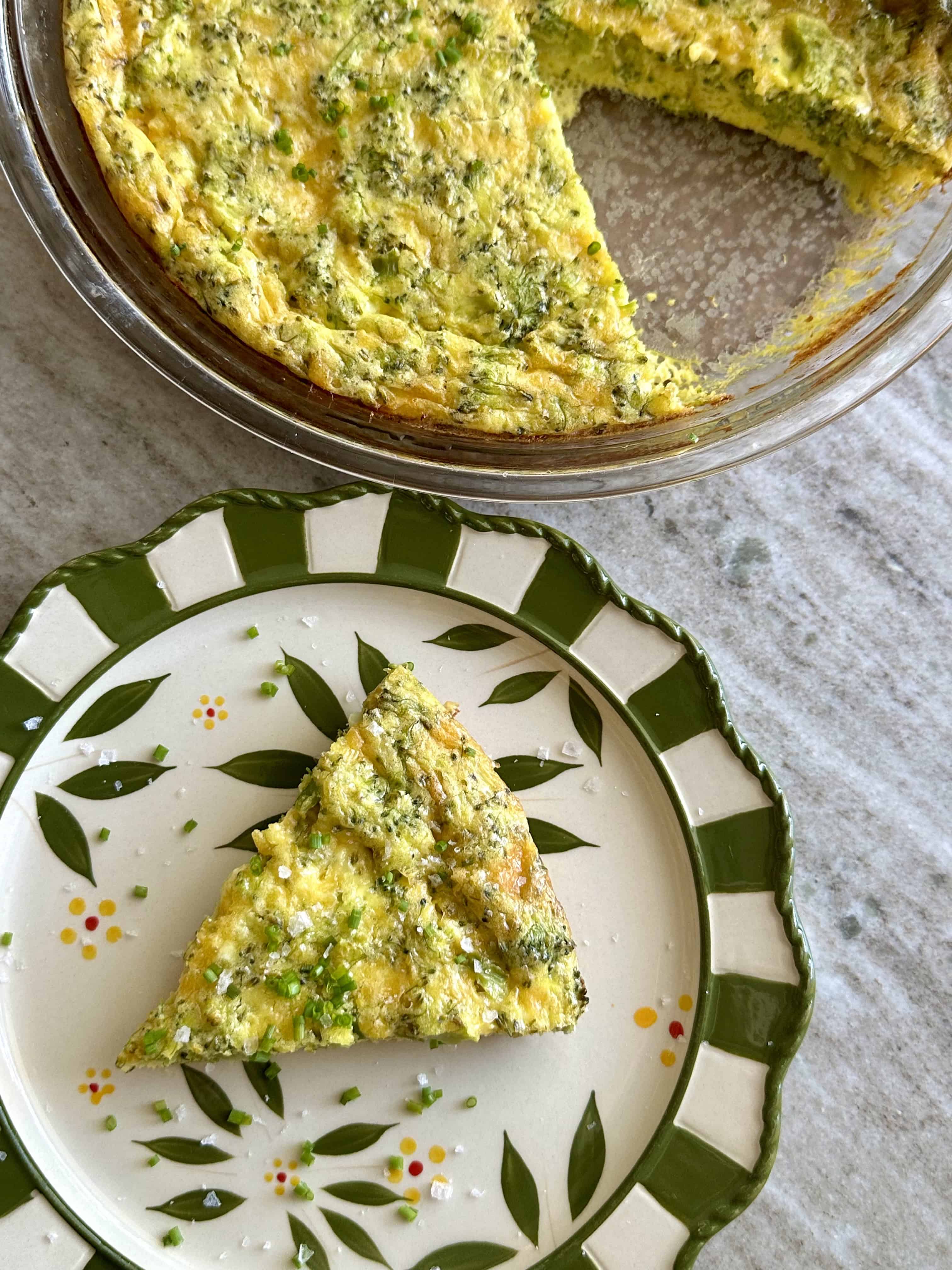 Slice of broccoli and cheese quiche on a floral green plate with a pie dish of quiche in the background.