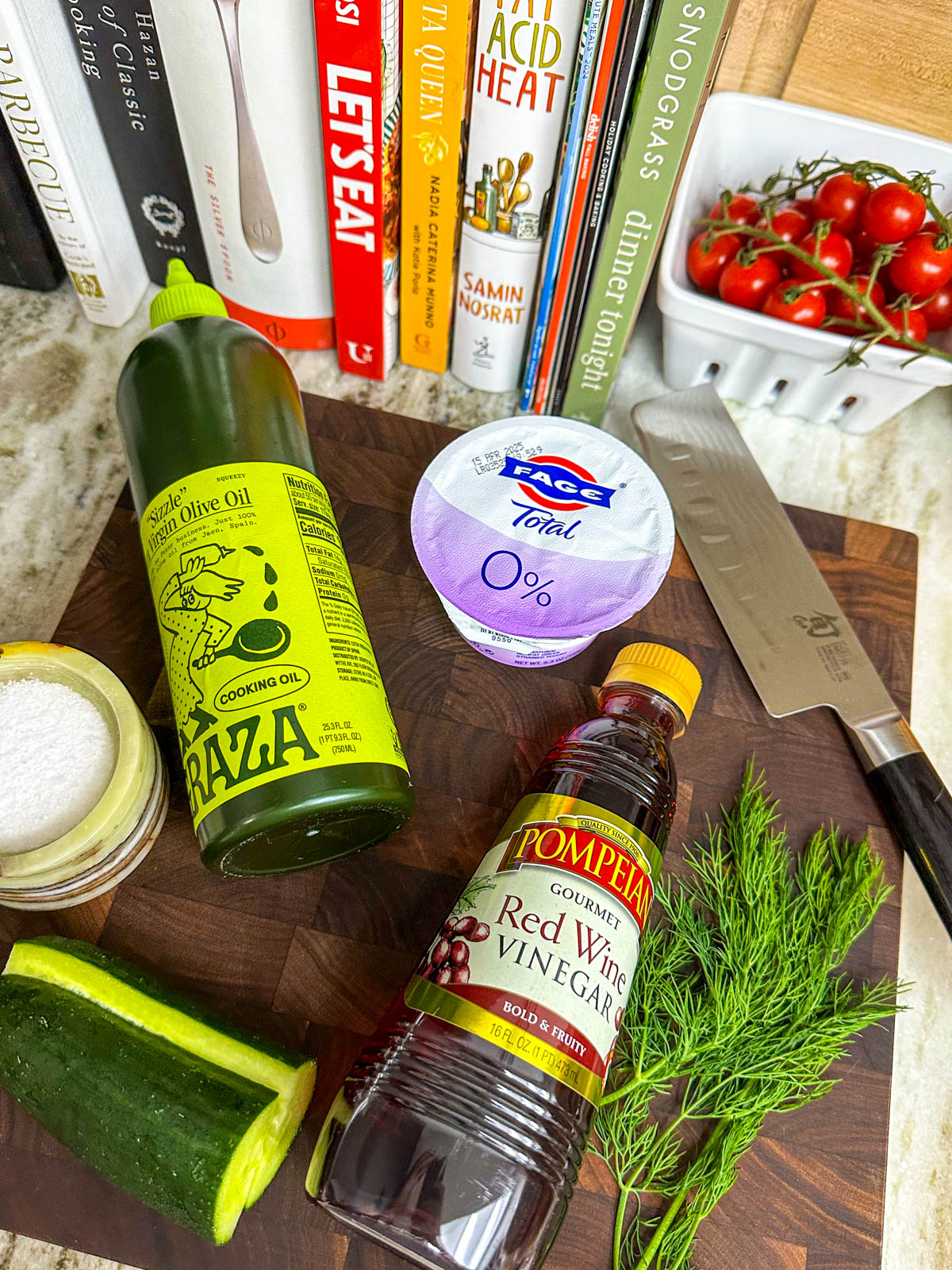 A bottle of olive oil, a cup of greek yogurt, a sprig of dill, half a cucumber, red wine vinegar, and salt on top of a cutting board. 