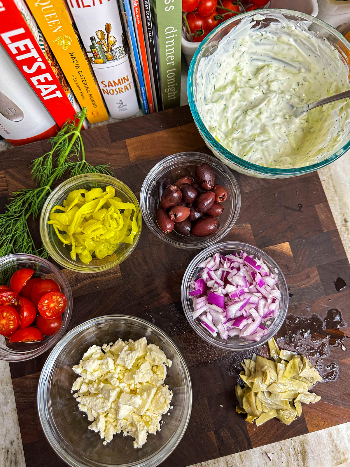 Bowls of toppings including feta, cherry tomatoes, red onion, pepperonicinis, Kalamata olives, tzatziki sauce, and marinated artichoke hearts on top of a cutting board. 
