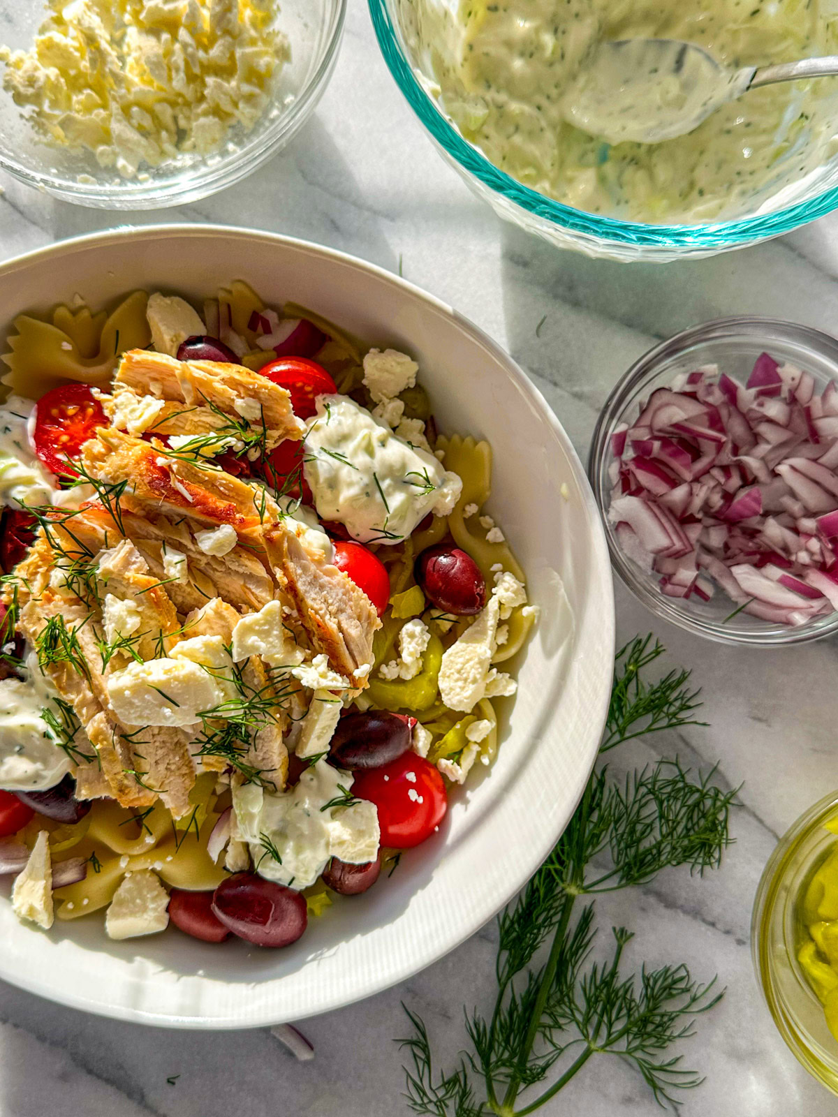 A white bowl filled with tzatziki sauce, pasta, chicken, cherry tomatoes, feta, chopped dill, pepperoncinis, Kalamata olives, marinated artichoke hearts with a bowl of feta and a sprig of dill in the background. 