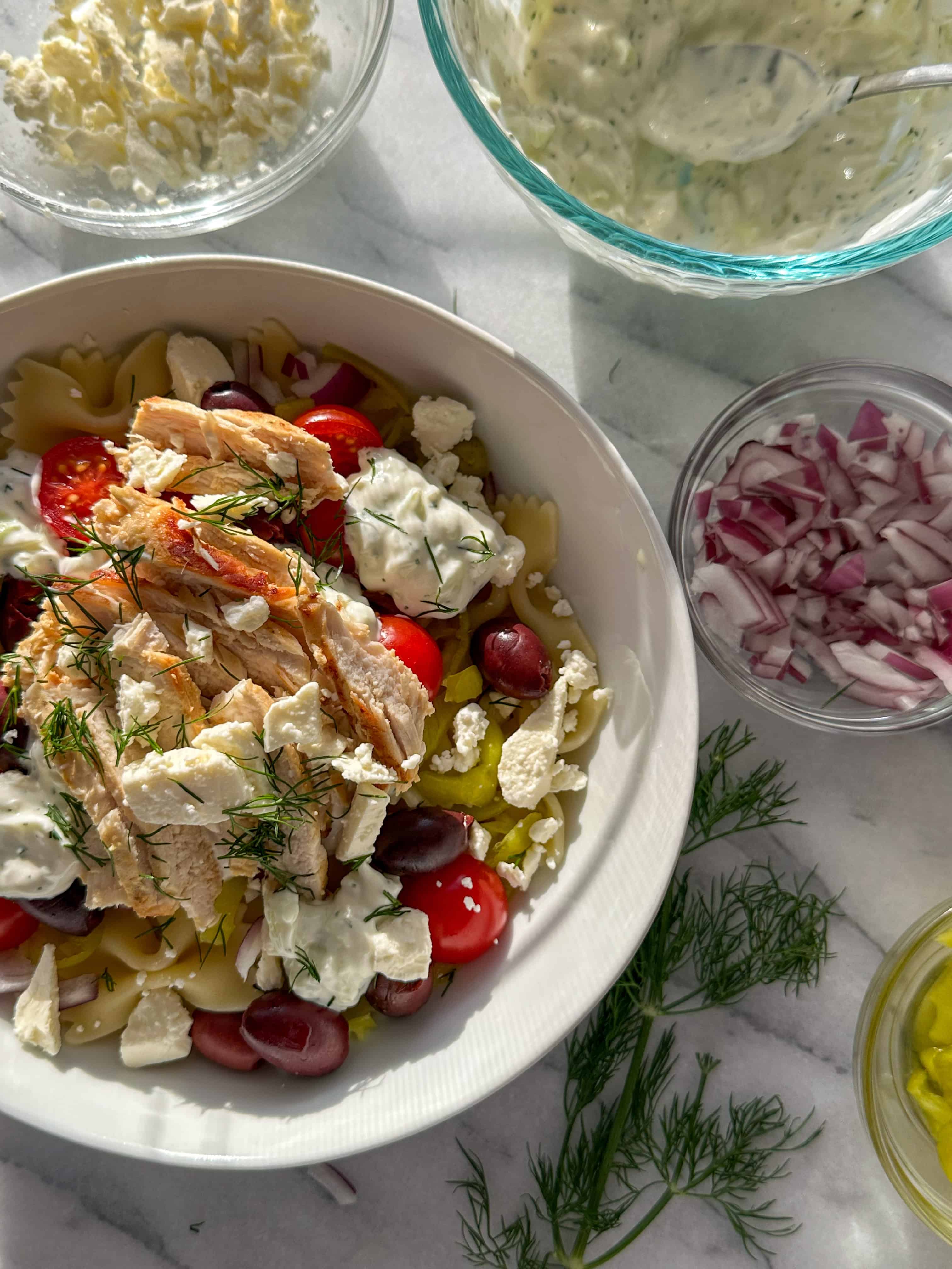 A white bowl filled with tzatziki sauce, pasta, chicken, cherry tomatoes, feta, chopped dill, pepperoncinis, Kalamata olives, marinated artichoke hearts with a bowl of feta and a sprig of dill in the background. 