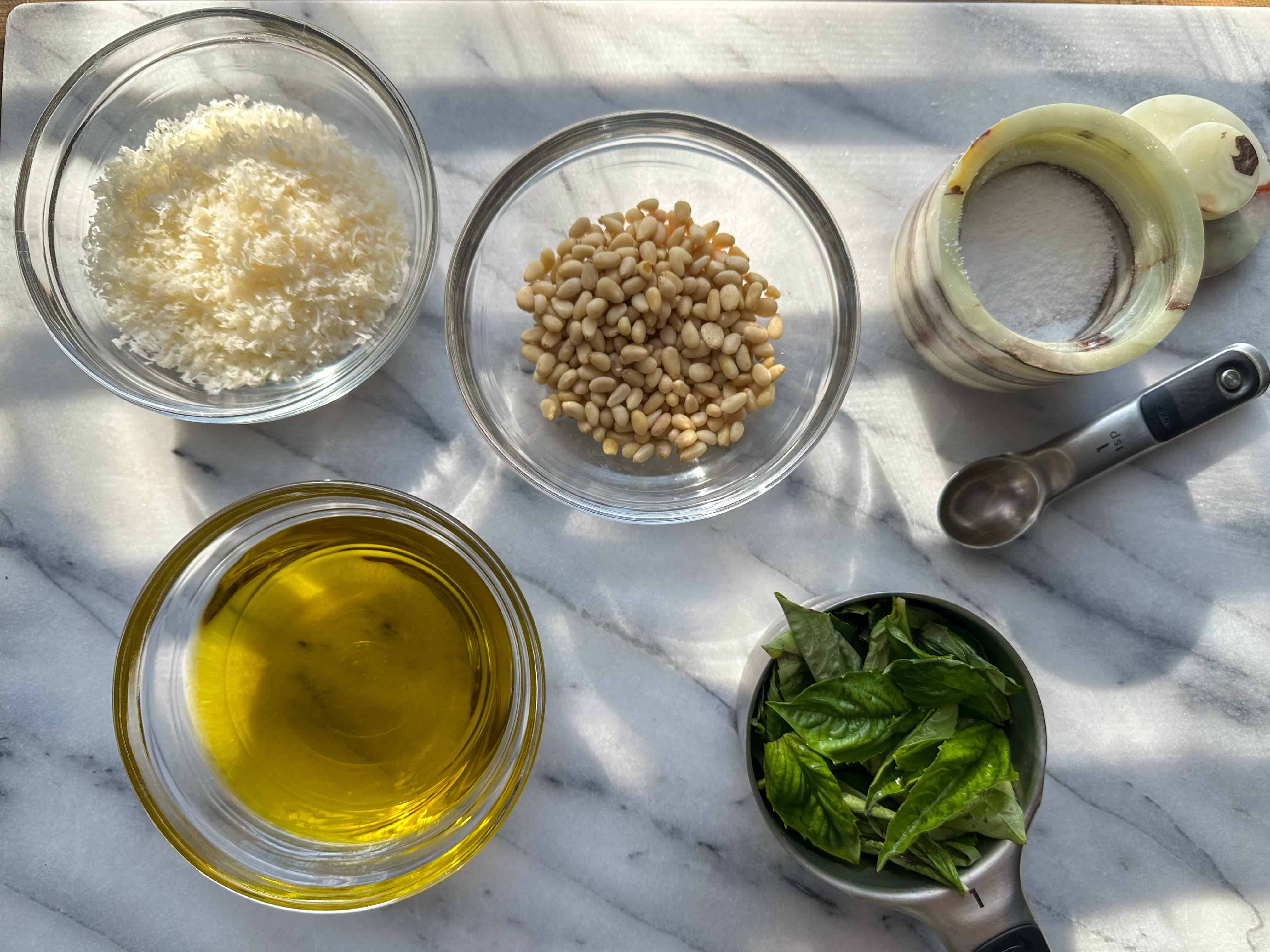 Clear ingredients bowls filled with parmigiano reggiano, pine nuts, basil, olive oil, and kosher salt.