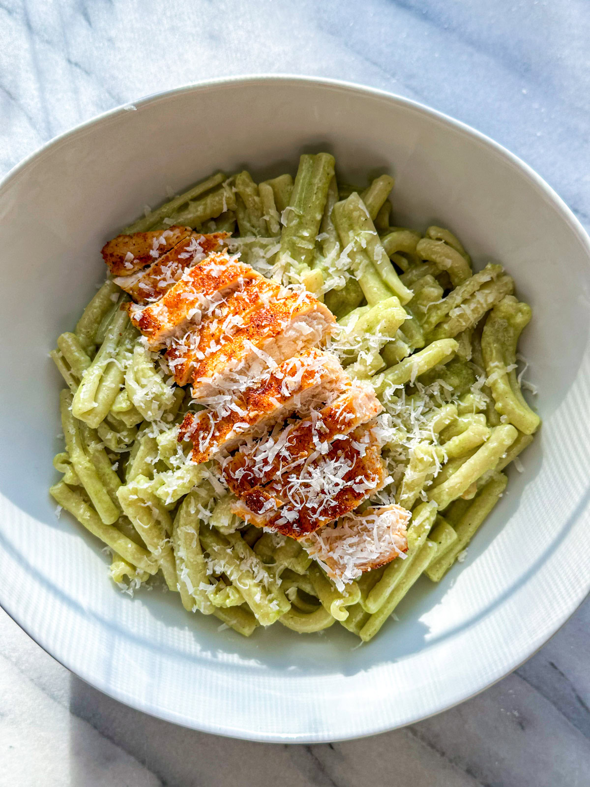 A white bowl filled with creamy pesto pasta topped with a sliced chicken breast and grated parmigiano reggiano. 