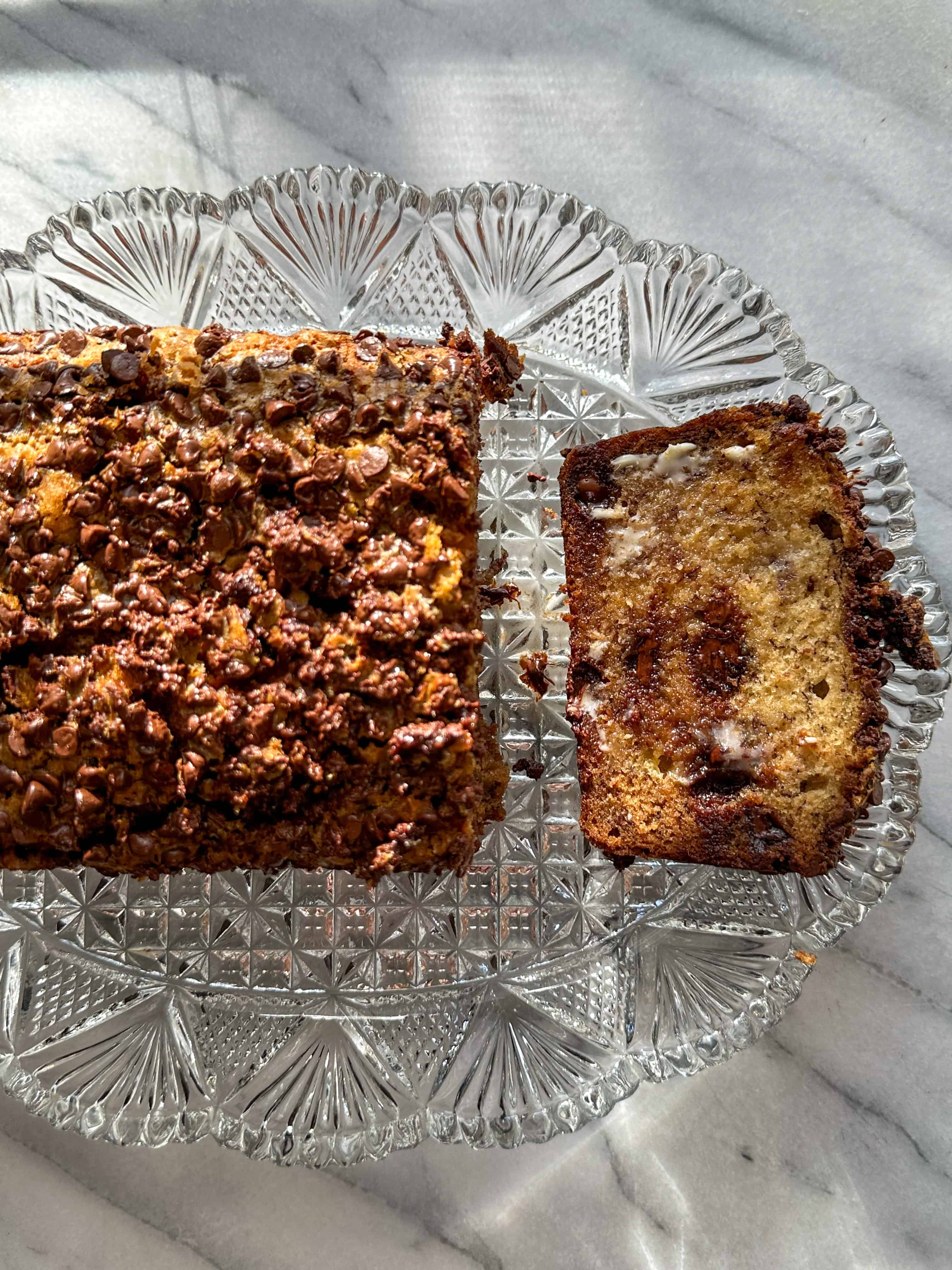 Loaf of banana bread topped with mini chocolate chips with a slice smeared with butter on a glass serving dish.