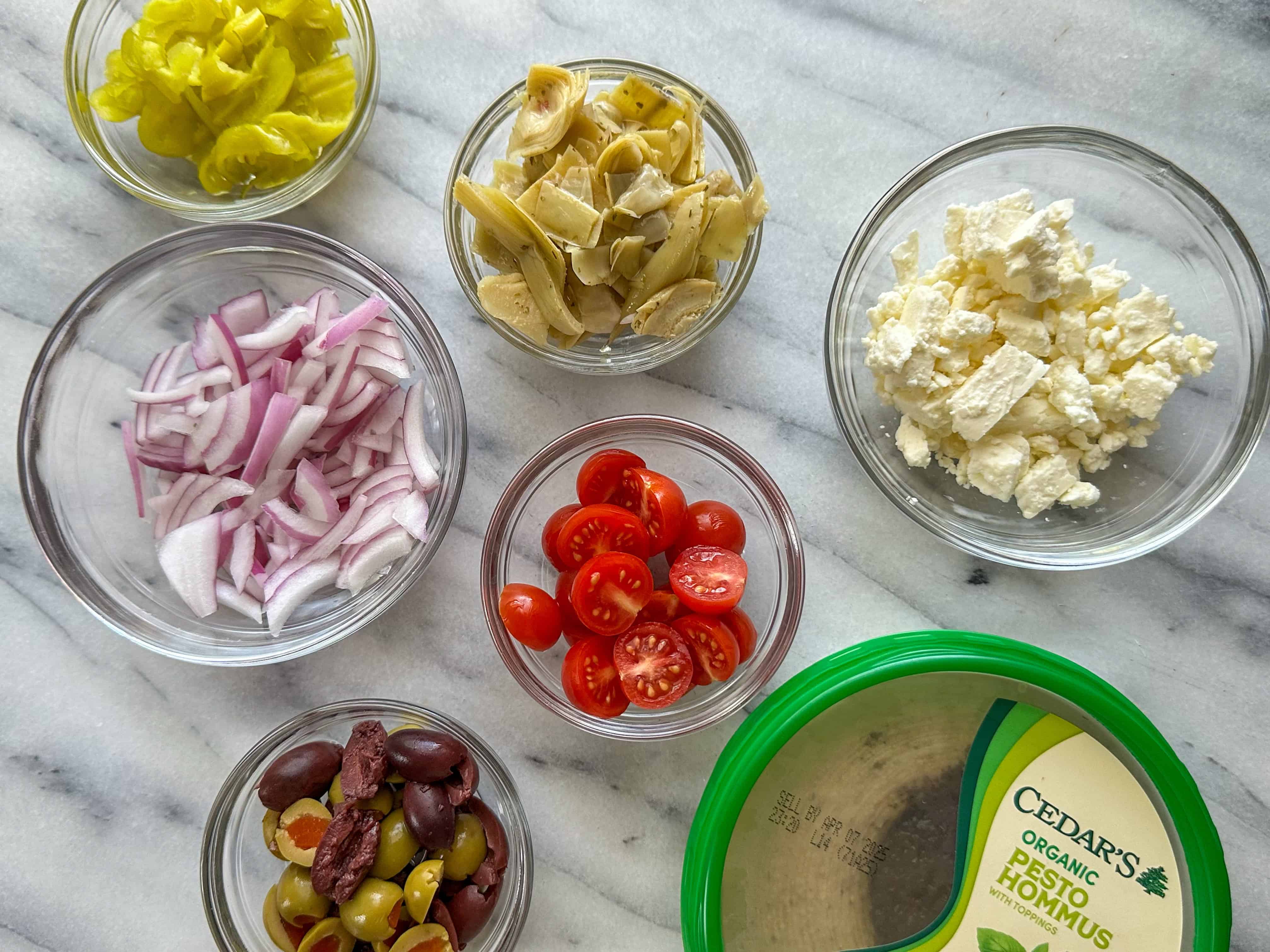 Clear bowls filled with peppers, marinated artichoke hearts, feta, chopped red onion, cherry tomatoes, green and Kalamata olives and a dish of hummus.