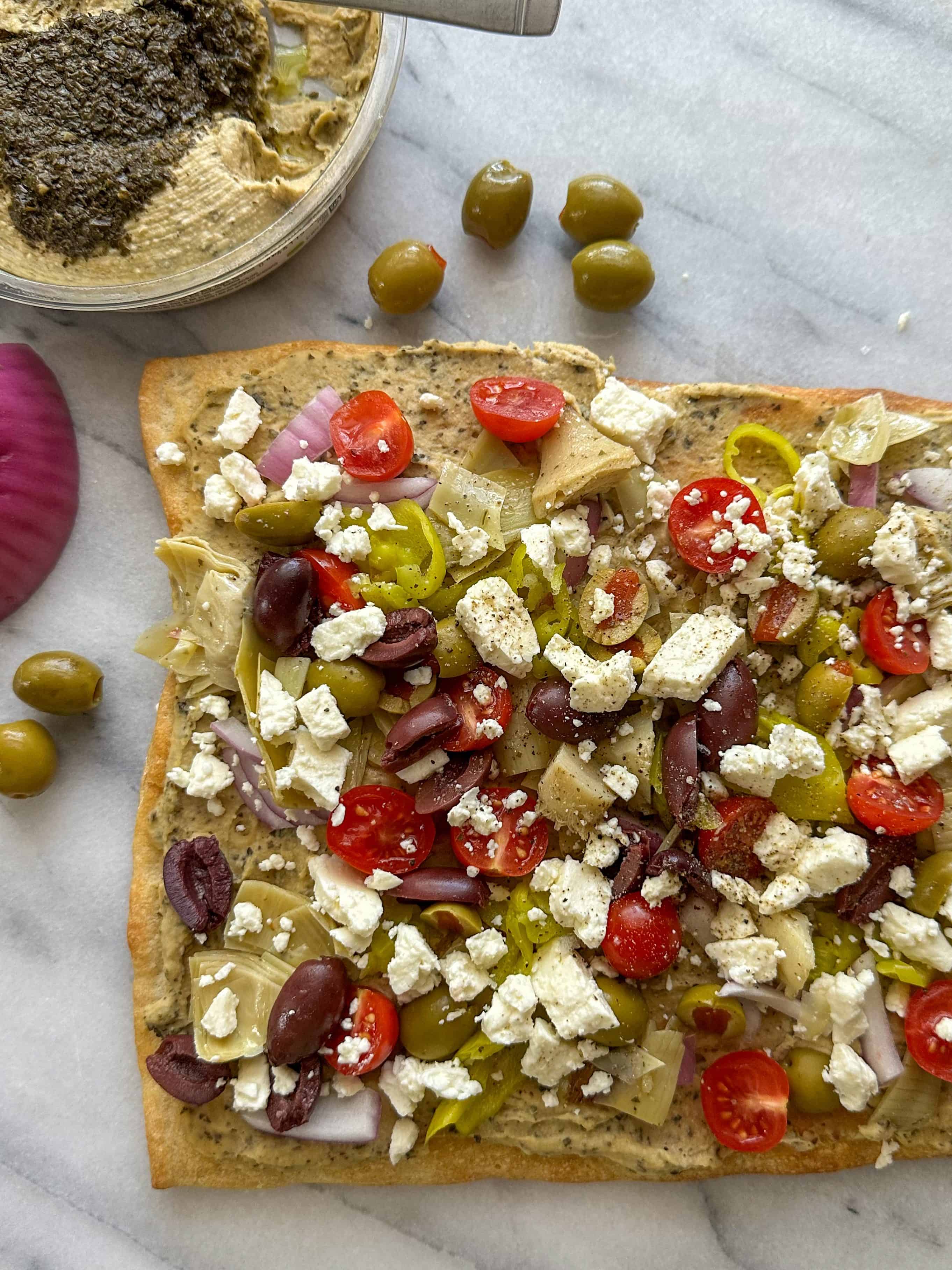 A mediterranean pizza sitting on a marble cutting board topped with hummus, feta, cherry tomatoes, olives, artichoke hearts, and red onion with loose olives in the background.