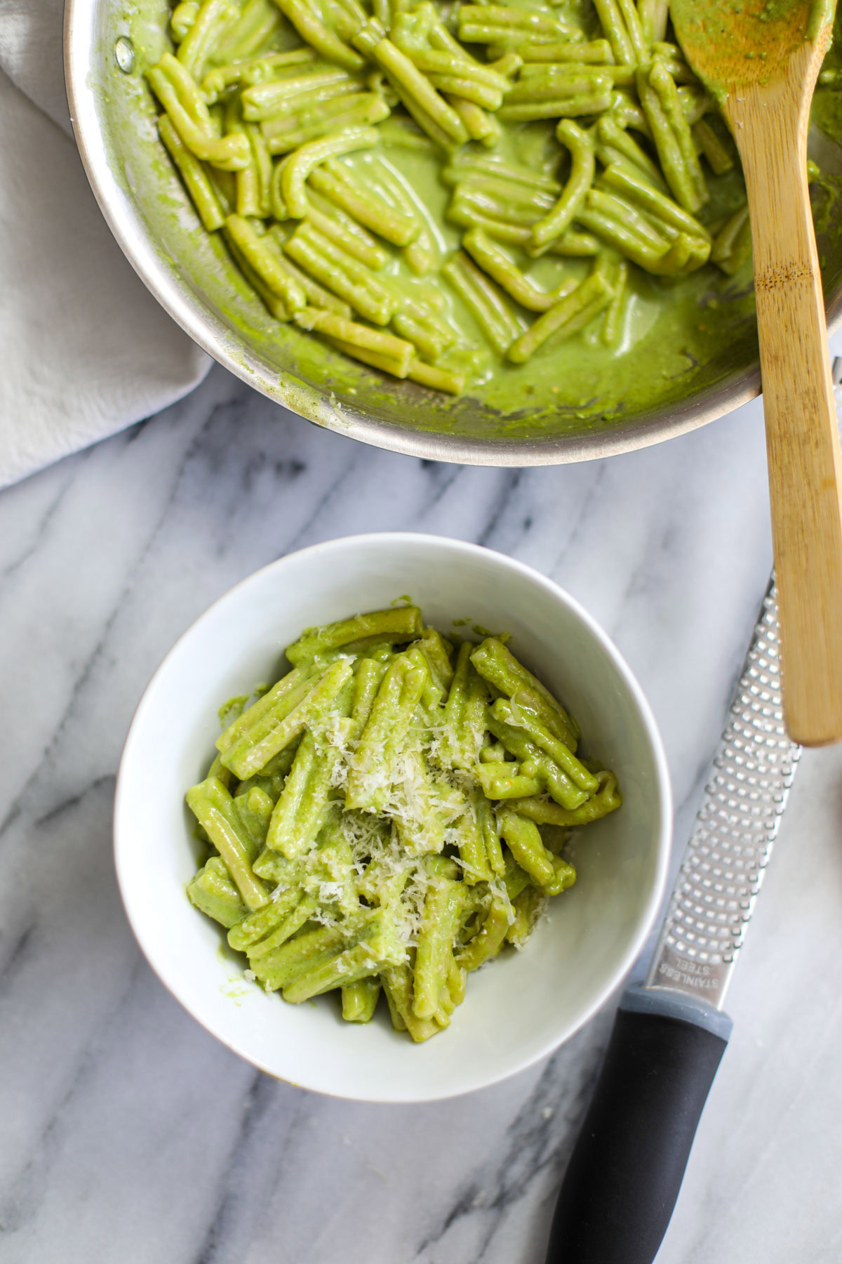 A white bowl of creamy pesto pasta next to the stainless pan full of pasta and a microplane grater. 