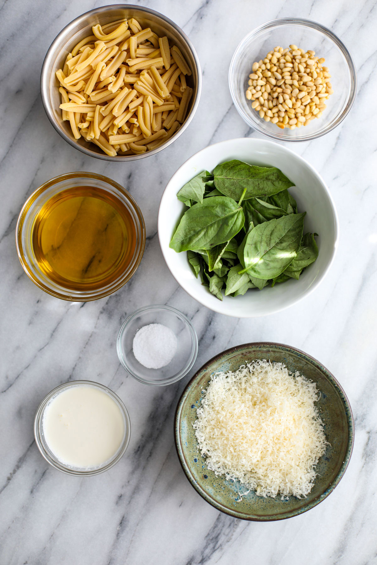 A stainless steel bowl of pasta, a glass bowl of pine nuts, glass bowls of heavy cream and salt, a blue bowl of parmigiana and a white bowl of basil.