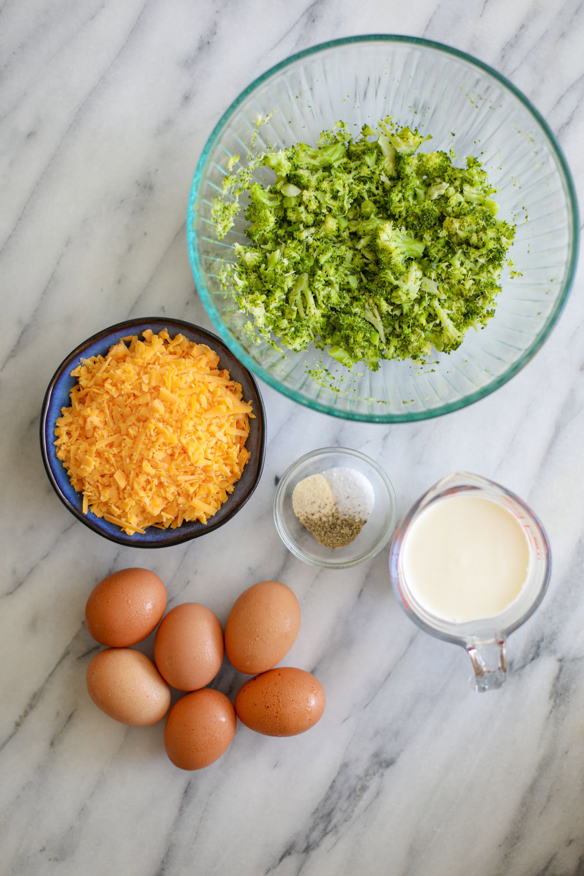 A large glass mixing bowl of chopped broccoli, a blue bowl of shredded cheese, six eggs, a small glass bowl of seasonings, and a measuring cup of milk. 