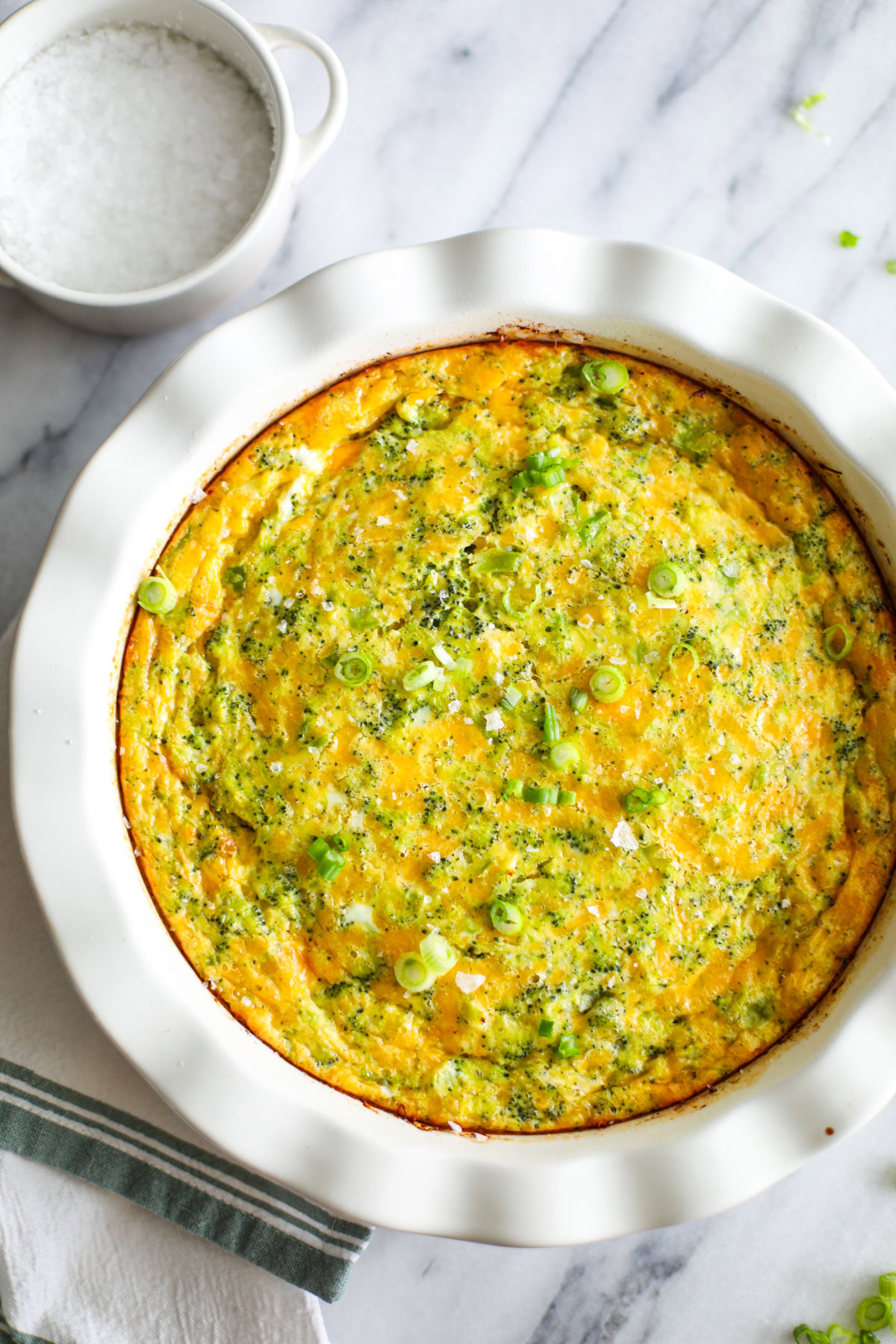 A broccoli cheese quiche in a white ruffled pie dish with a ramekin of flaky salt and white and green dish towel off to the side. 