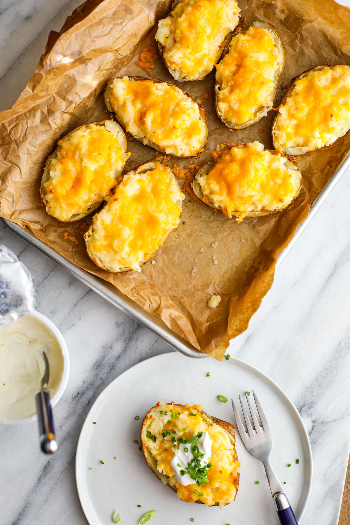 A tray of twice baked potatoes next to a white plate with a chive and sour cream topped potato and a blue fork.