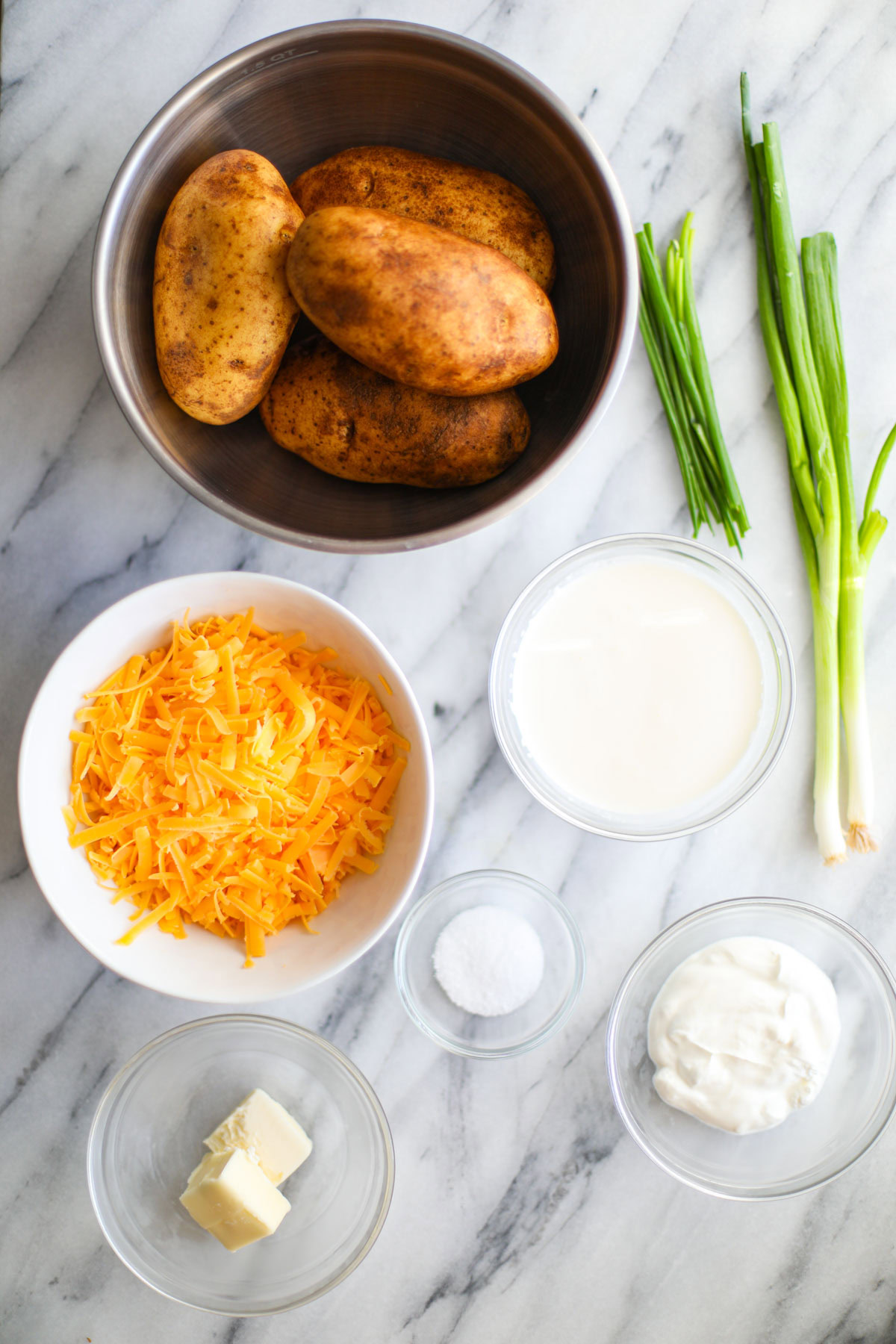 A stainless steel bowl of potatoes, chives, green onions, bowls of cream, sour cream, cheese, salt, and butter on a marble counter.