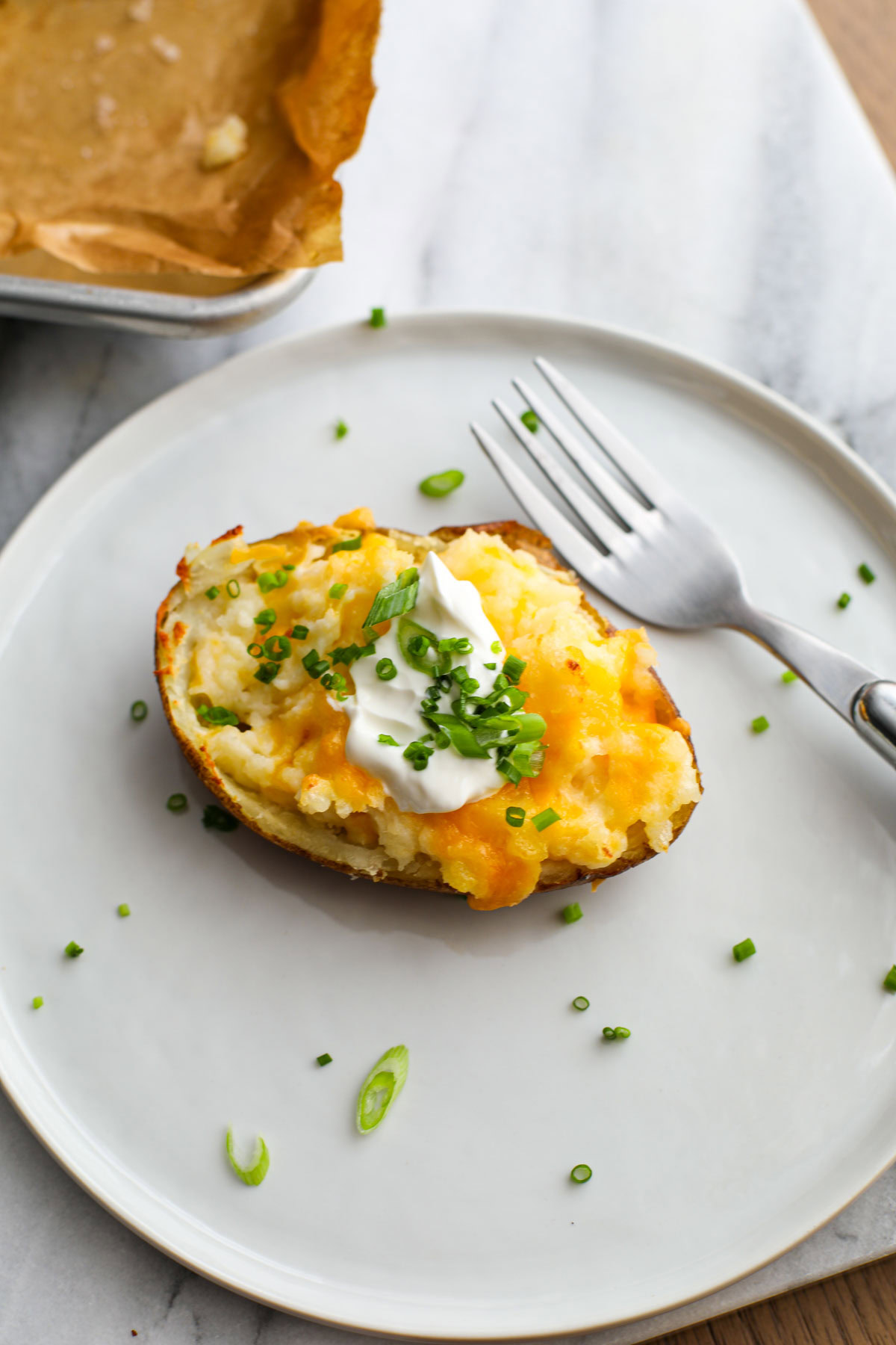 A twice baked potato topped with sour cream, chives, and green onions on a white plate with a stainless fork.