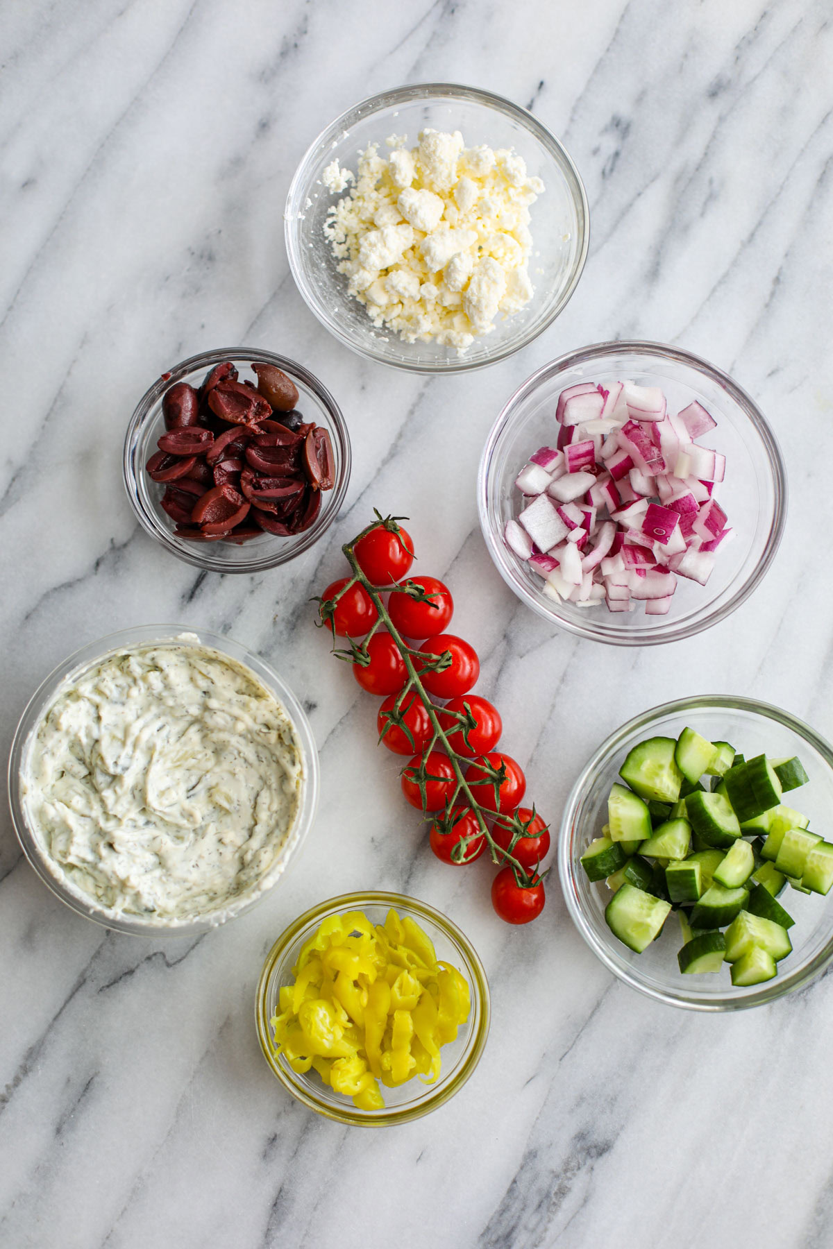 Glass bowls of crumbled feta, sliced Kalamata olives, sliced red onion, cucumber, and pepperonicinis, cherry tomatoes attached to a vine, and bowl of tzatziki sauce.
