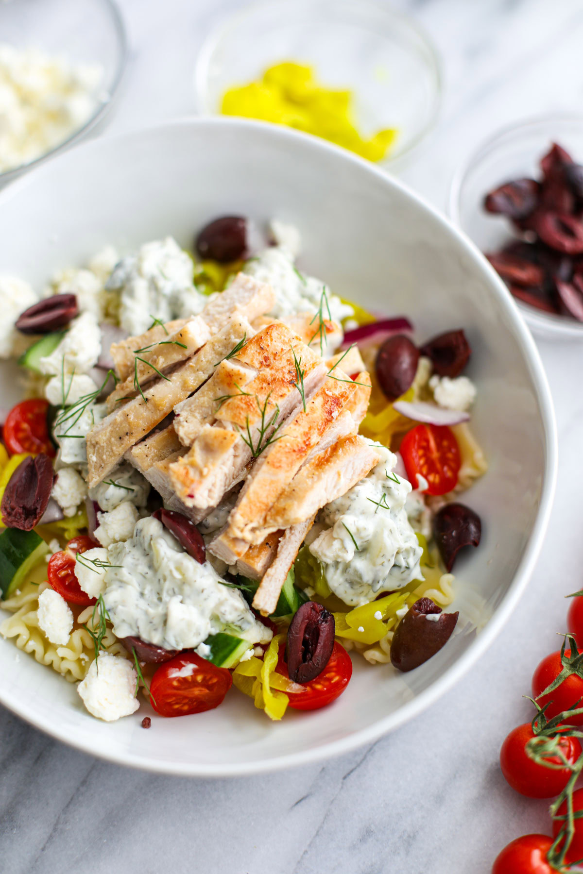 A white bowl filled with tzatziki sauce, pasta, chicken, cherry tomatoes, feta, chopped dill, pepperoncinis, Kalamata olives, marinated artichoke hearts with a bowl of feta and a sprig of dill in the background. 
