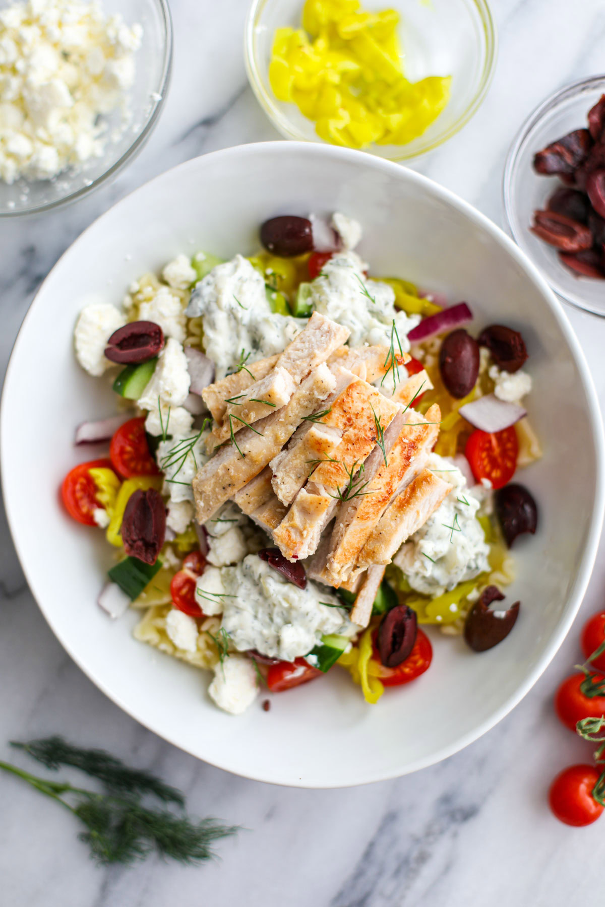 A white bowl filled with tzatziki sauce, pasta, chicken, cherry tomatoes, feta, chopped dill, pepperoncinis, Kalamata olives, marinated artichoke hearts with a bowl of feta and a sprig of dill in the background. 