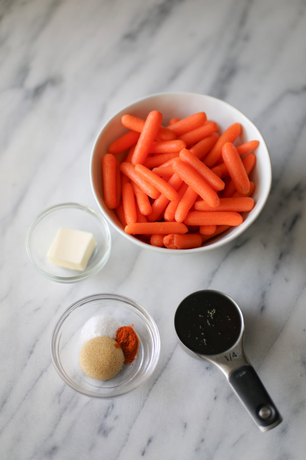 A white bowl filled with baby carrots, a clear bowl with a tablespoon of butter, a clear bowl with a tablespoon of brown sugar with salt and cayenne pepper, and a 1/4 cup measuring cup filled with honey.