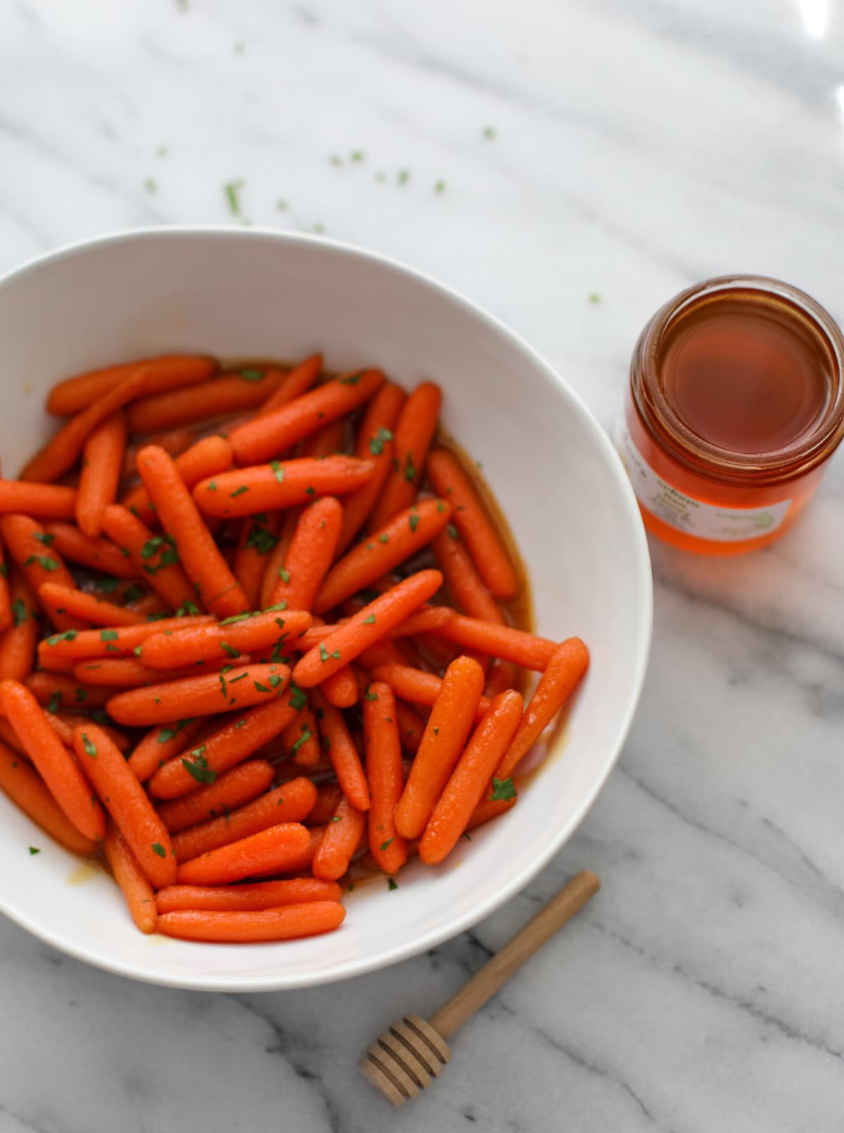 A white bowl filled with baby carrots in a hot honey glaze topped with chopped parsley.