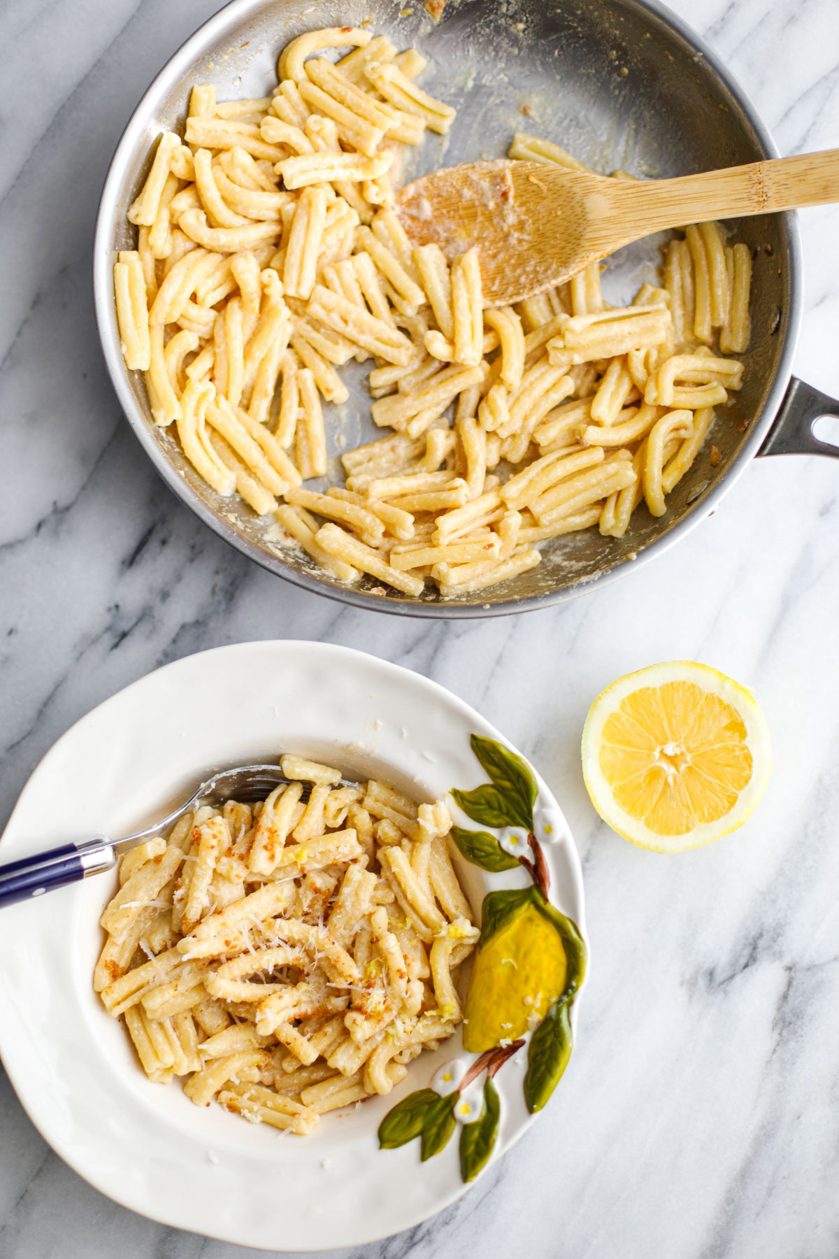 A white bowl of creamy lemon pasta with a blue fork next to a stainless steel pan of pasta and half a lemon beside it.