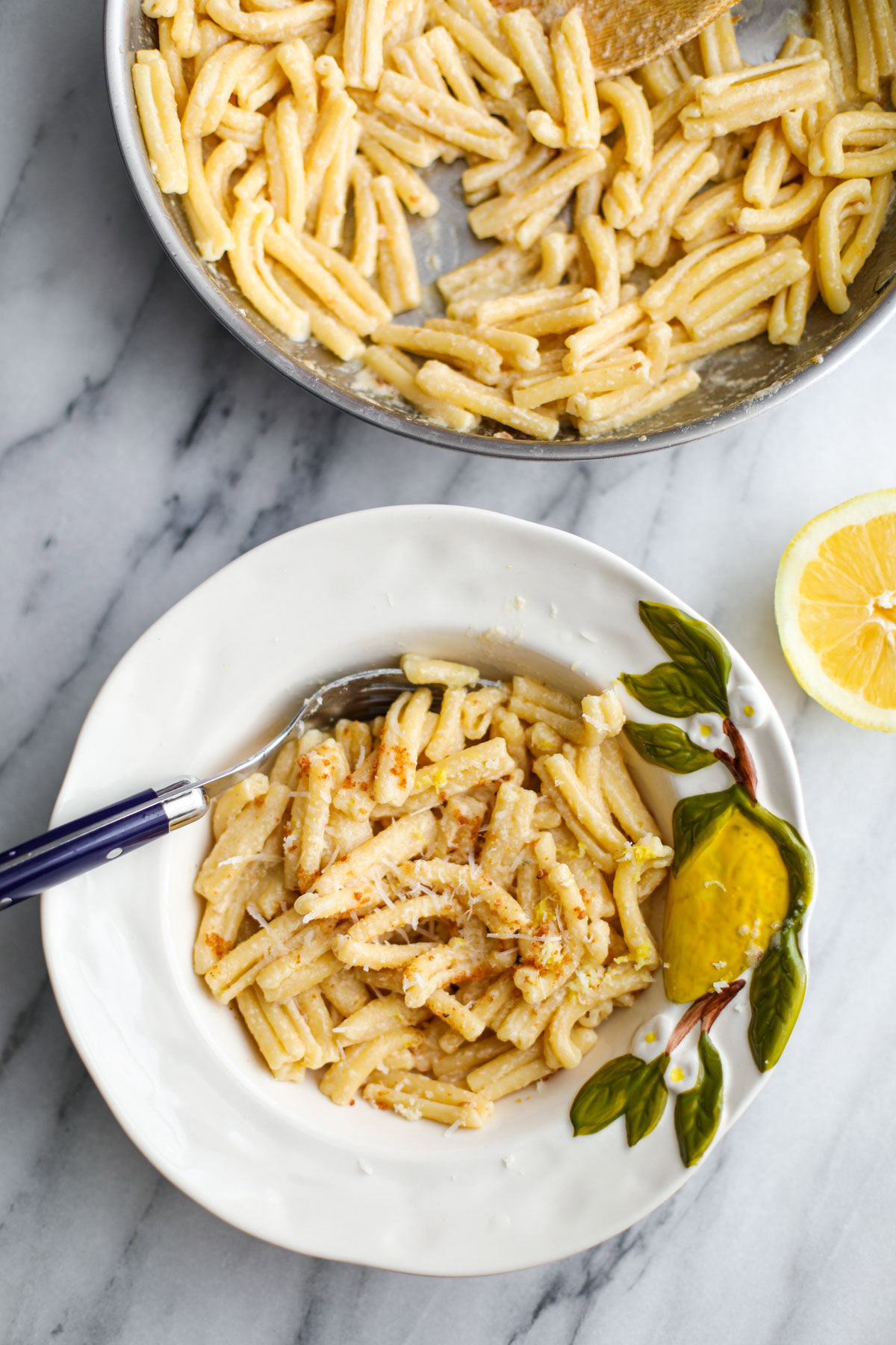 A white bowl of creamy lemon pasta with a blue fork next to a stainless steel pan of pasta and half a lemon beside it.