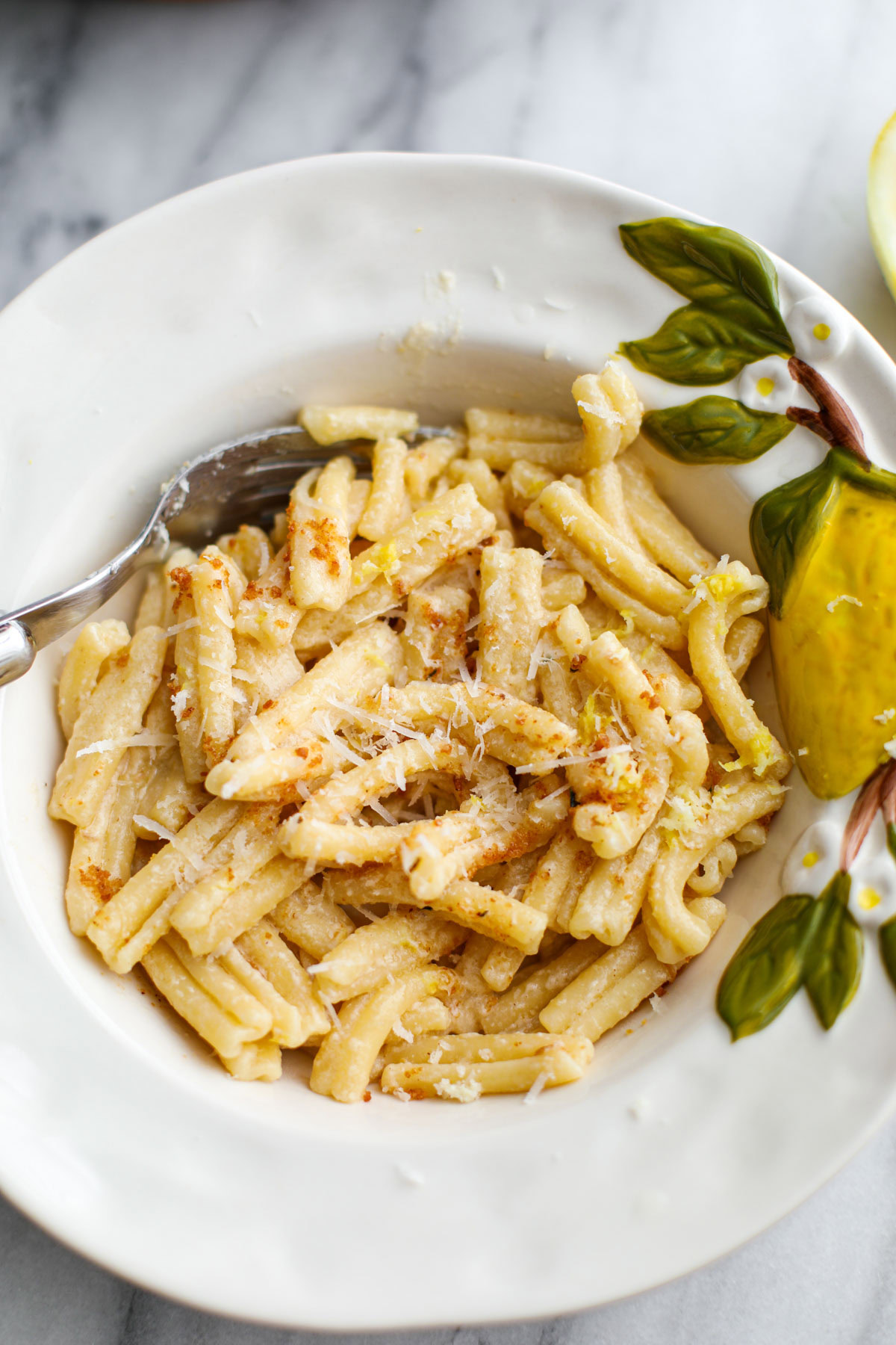 A white bowl with a lemon on it filled with pasta and topped with grated cheese and breadcrumbs.