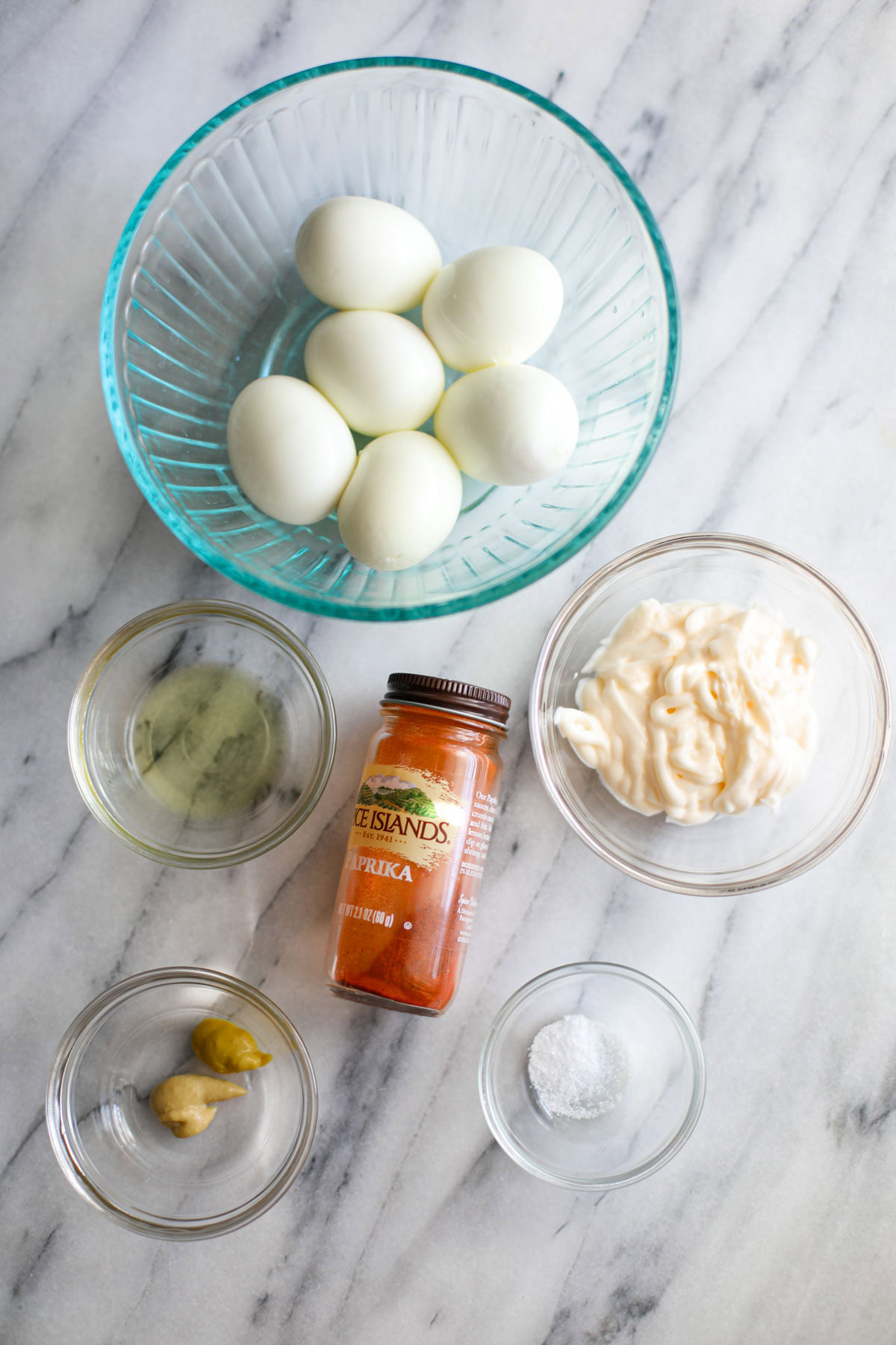 A clear bowl of peeled, hardboiled eggs, clear bowls of mayonnaise, mustard, pickle juice, and salt next to a bottle of paprika.