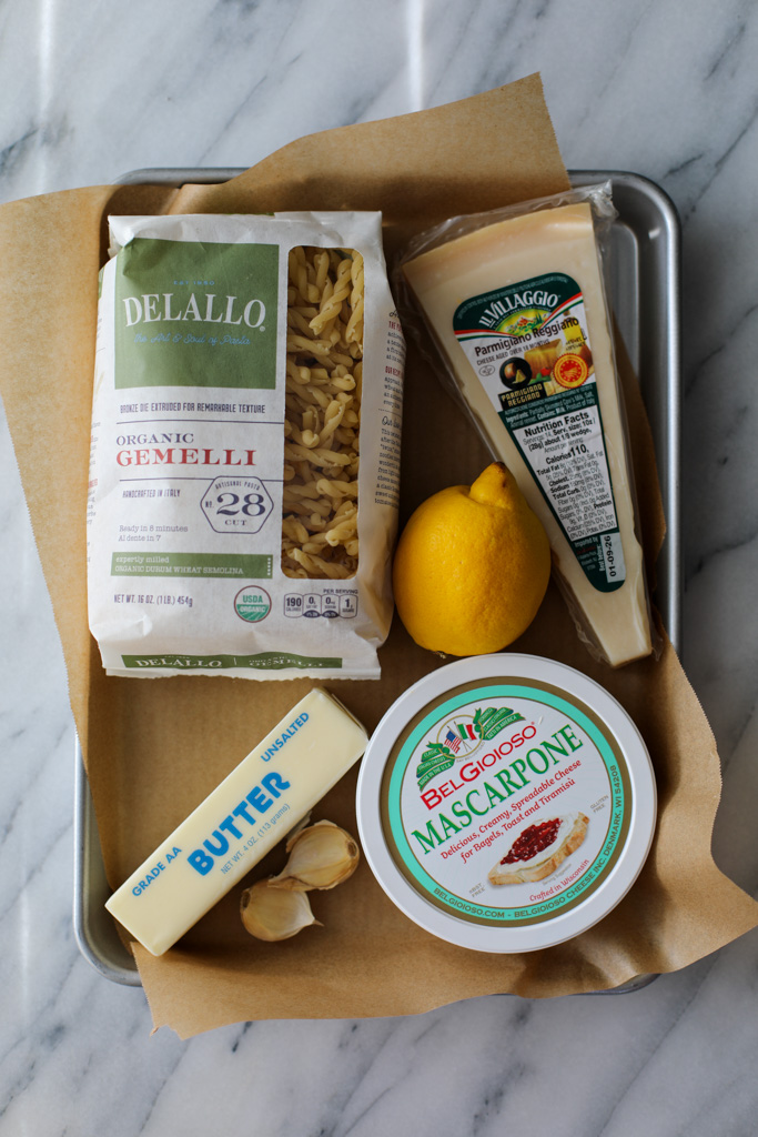 A silver sheet pan covered with brown parchment paper with a bag of gemelli pasta, a lemon, a stick of butter, 2 cloves of garlic, marscapone, and a wedge of Parmigiano Reggiano.