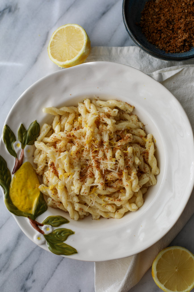 A white bowl with a painted lemon on it filled with creamy lemon pasta with half lemons in the background.