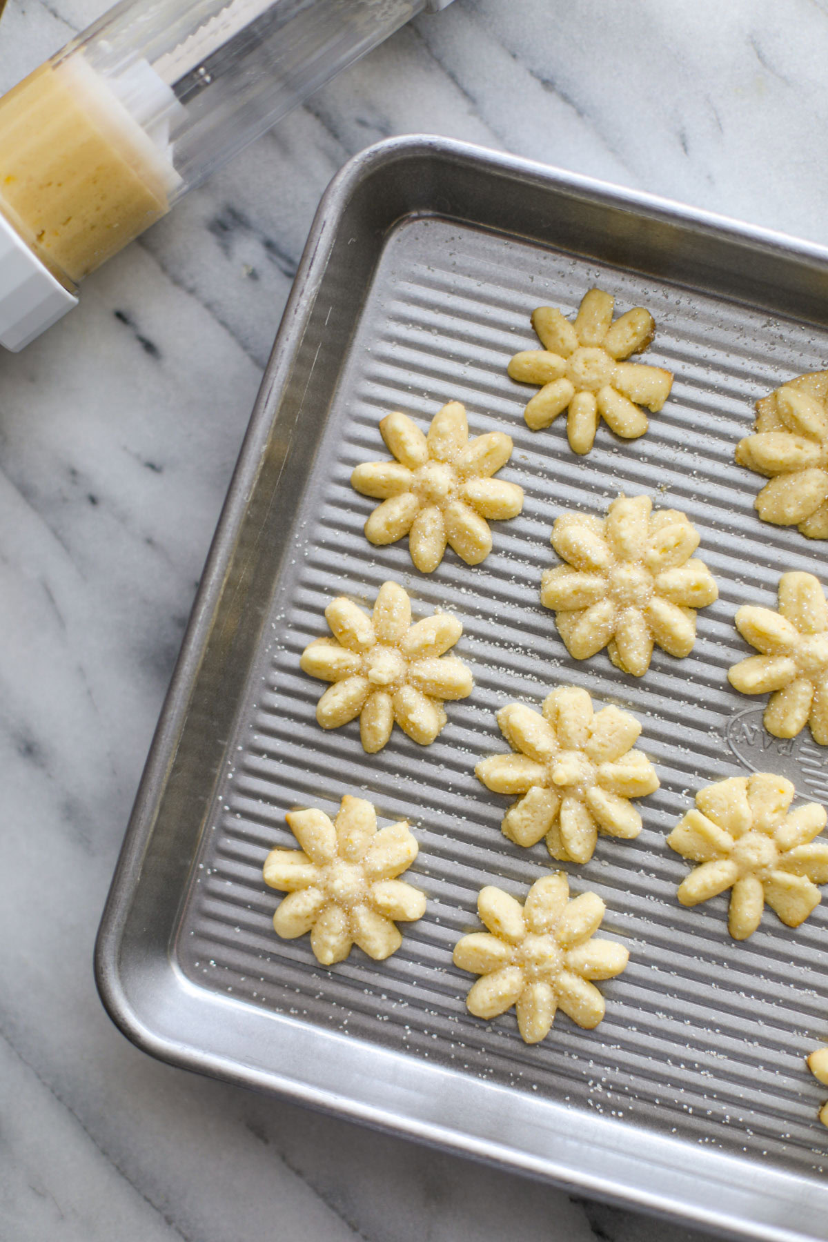 Flower shaped Lemon Spritz Cookies on a metal cookie sheet with sliced lemons and a cookie press in the background.