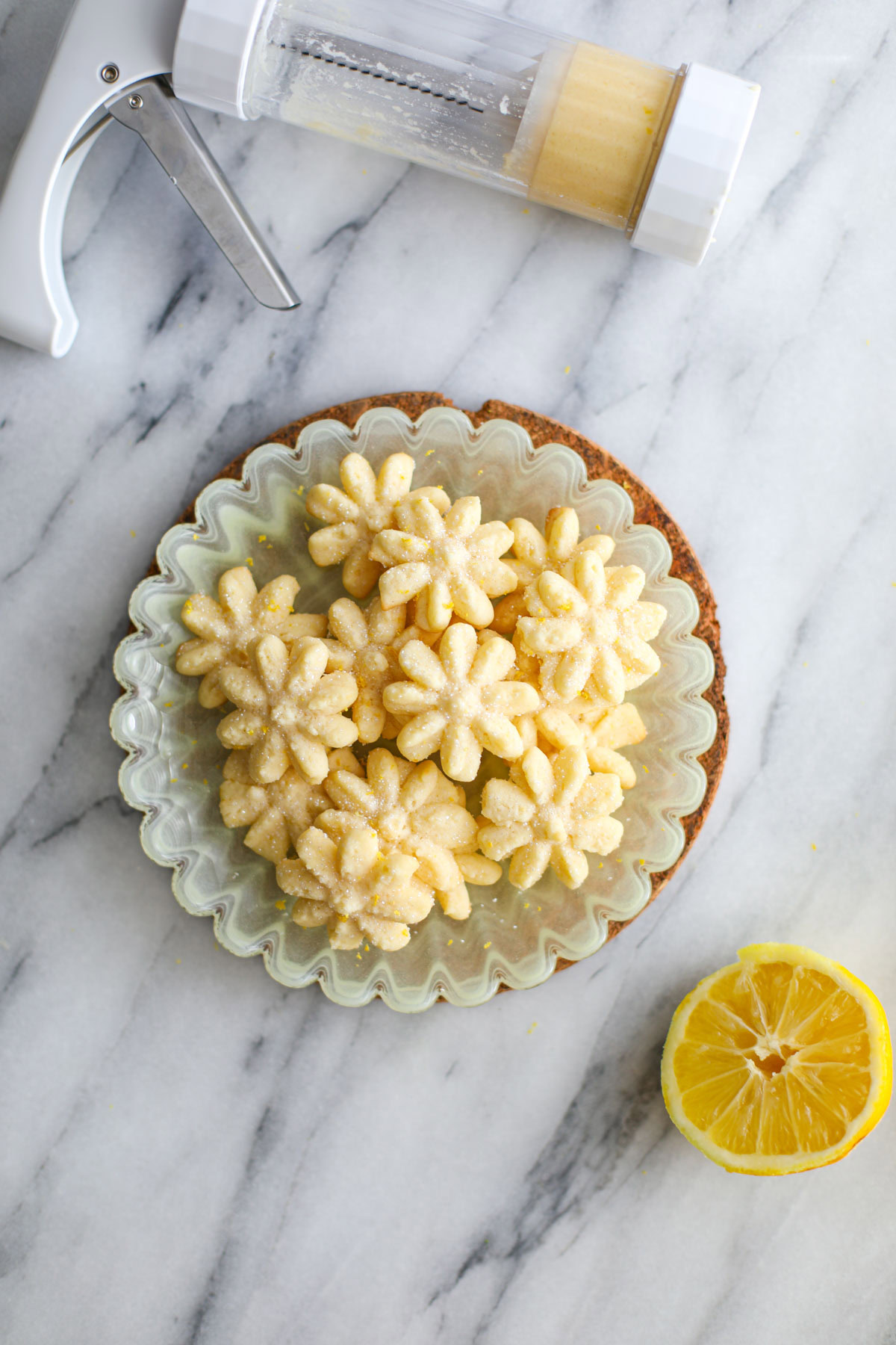 Flower shaped Lemon Spritz Cookies on a yellow plate with a cookie press and half a lemon next to it. 