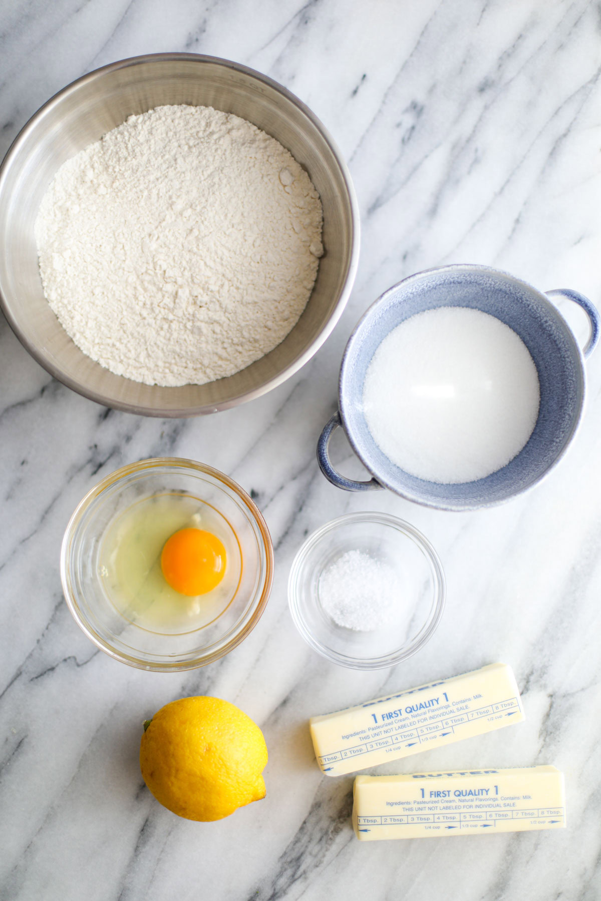 A stainless steel bowl of flour, a blu bowl of sugar, an egg, a glass bowl of salt, 2 sticks of butter and a whole lemon. 