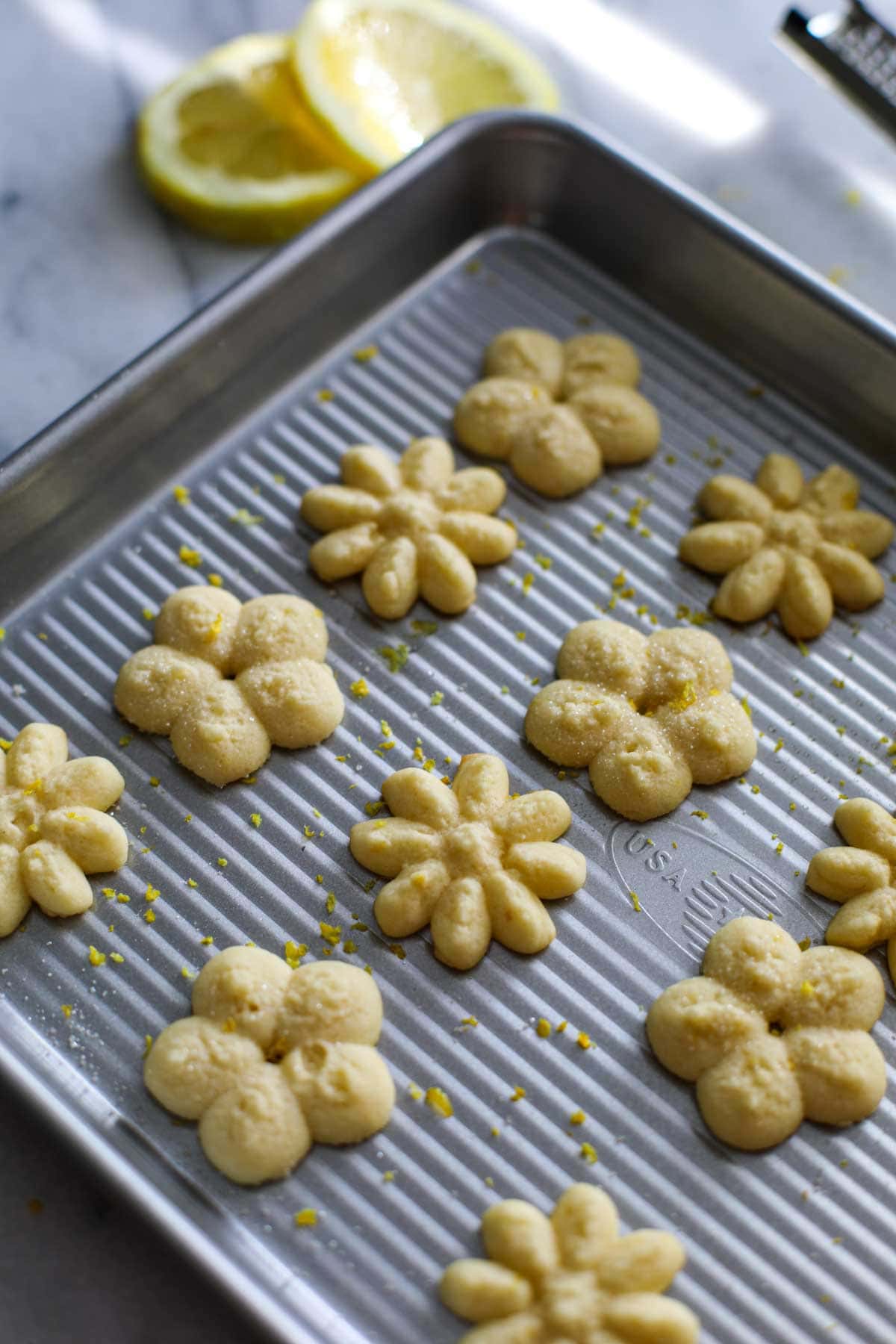 Flower shaped Lemon Spritz Cookies on a silver cookie sheet with sliced lemons in the background.