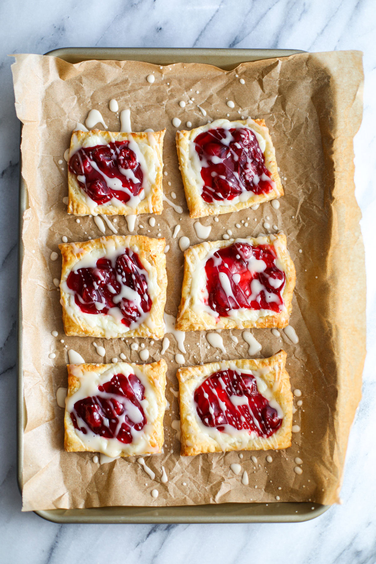 Brown parchment paper topped with square cherry cheese danishes drizzled with a white glaze with a bowl of cherry pie filling in the background.