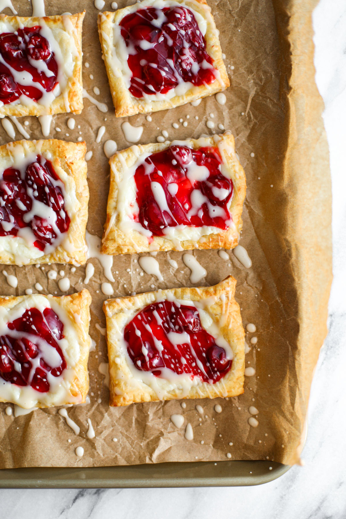 Brown parchment paper topped with square cherry cheese danishes drizzled with a white glaze with a bowl of cherry pie filling in the background.