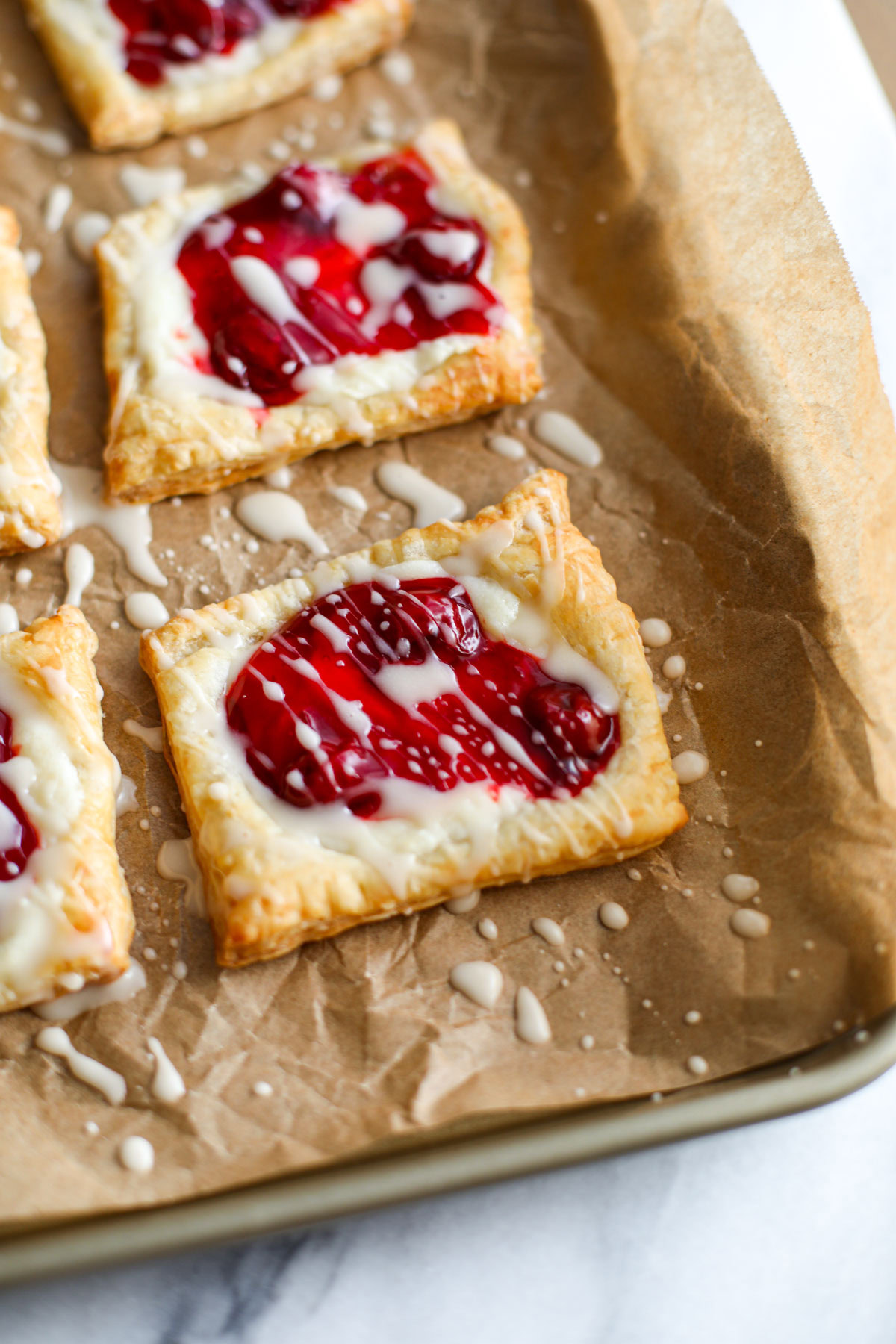 A cherry cheese danish drizzled with a white glaze on a gold sheet pan.