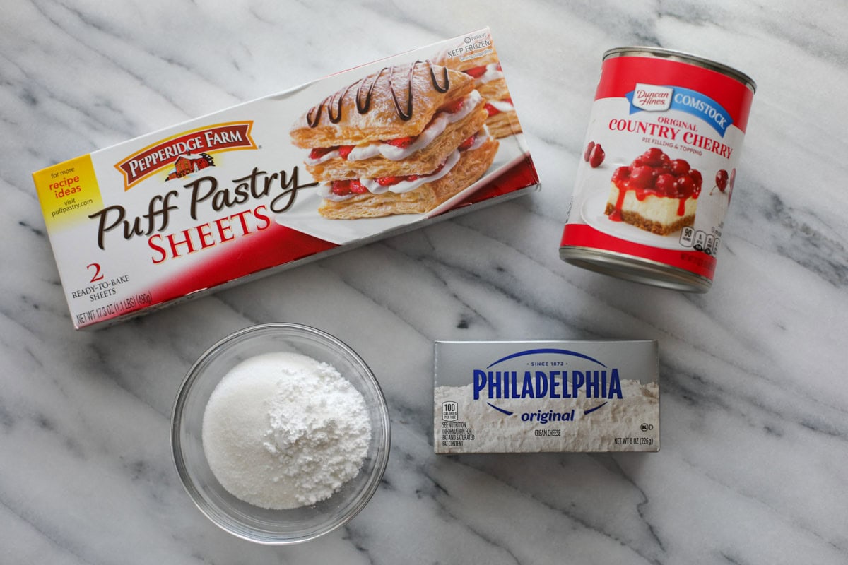 A box of Pepperidge Farm Puff Pastry sheets, a can of Duncan Hines country cherry pie filling, a bowl of white sugar, and a box of Philadelphia cream cheese on a marble table.