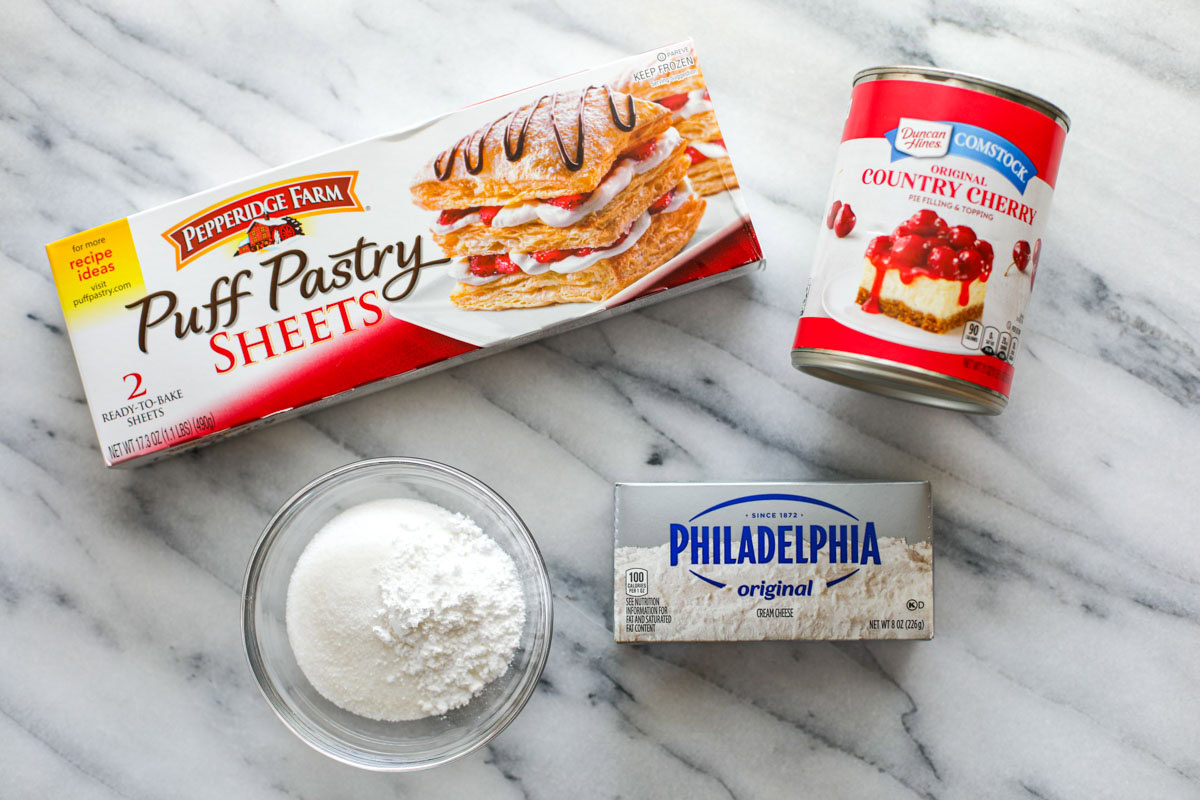 A box of Pepperidge Farm Puff Pastry sheets, a can of Duncan Hines country cherry pie filling, a bowl of white sugar, and a box of Philadelphia cream cheese on a marble table.
