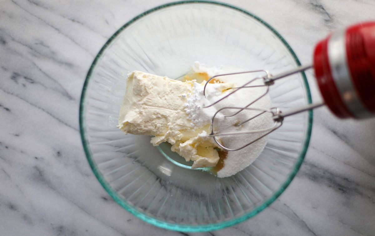 A clear mixing bowl containing a block of cream cheese, white sugar, vanilla extract, lemon juice and powdered sugar with a red hand mixer having off the edge of the bowl.