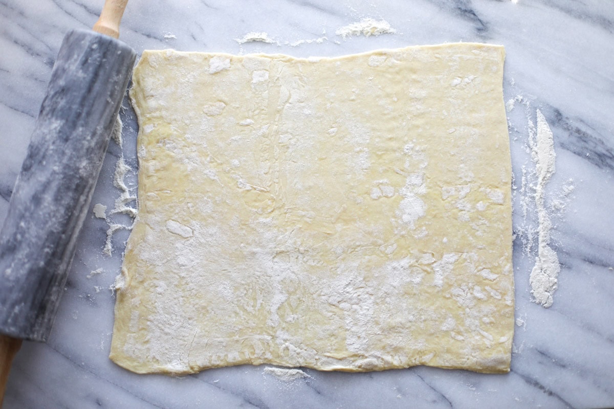 A large rectangle of rolled out puff pastry sheet with a marble roller on a marble board.