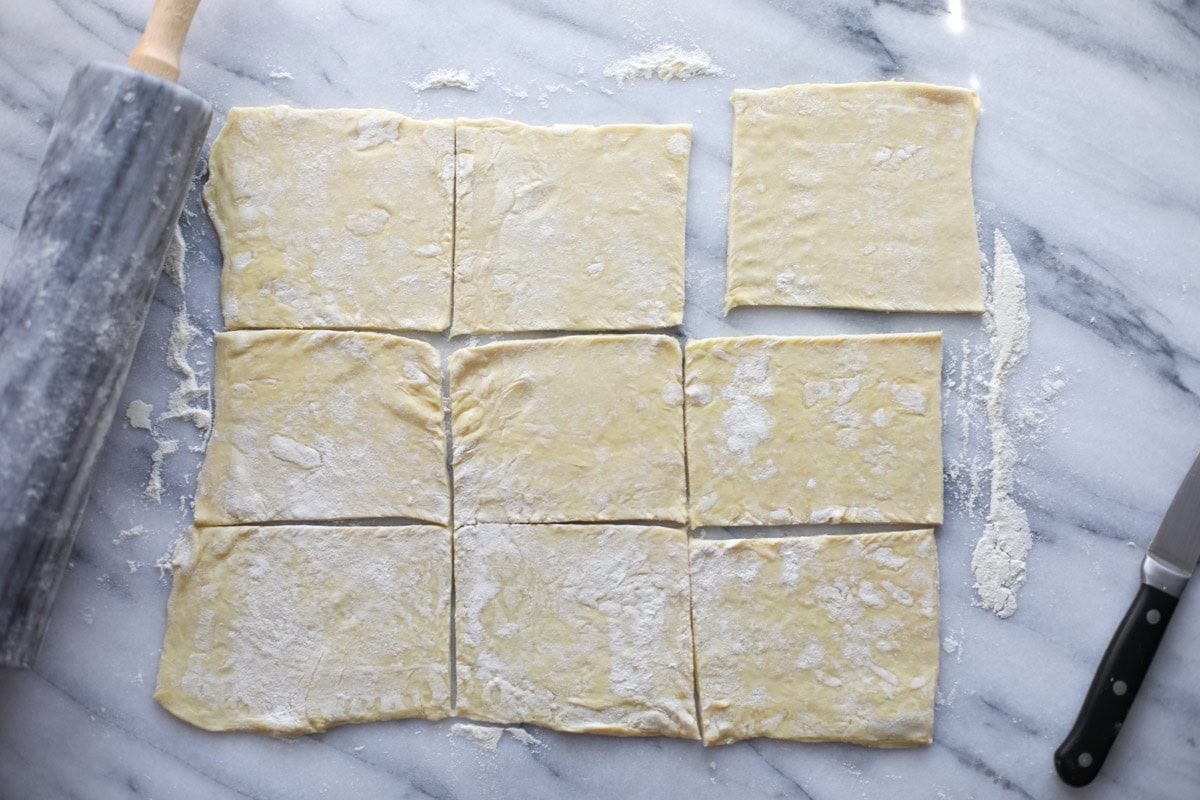 A large rectangle of rolled out puff pastry sheet cut into nine even squares with a marble roller on a marble board.
