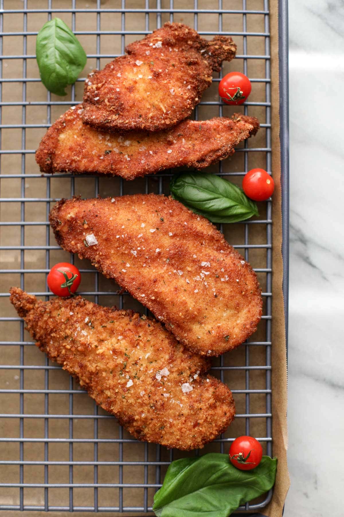 Four breaded and fried chicken cutlets resting on a cookie sheet with basil and cherry tomatoes surrounding them.