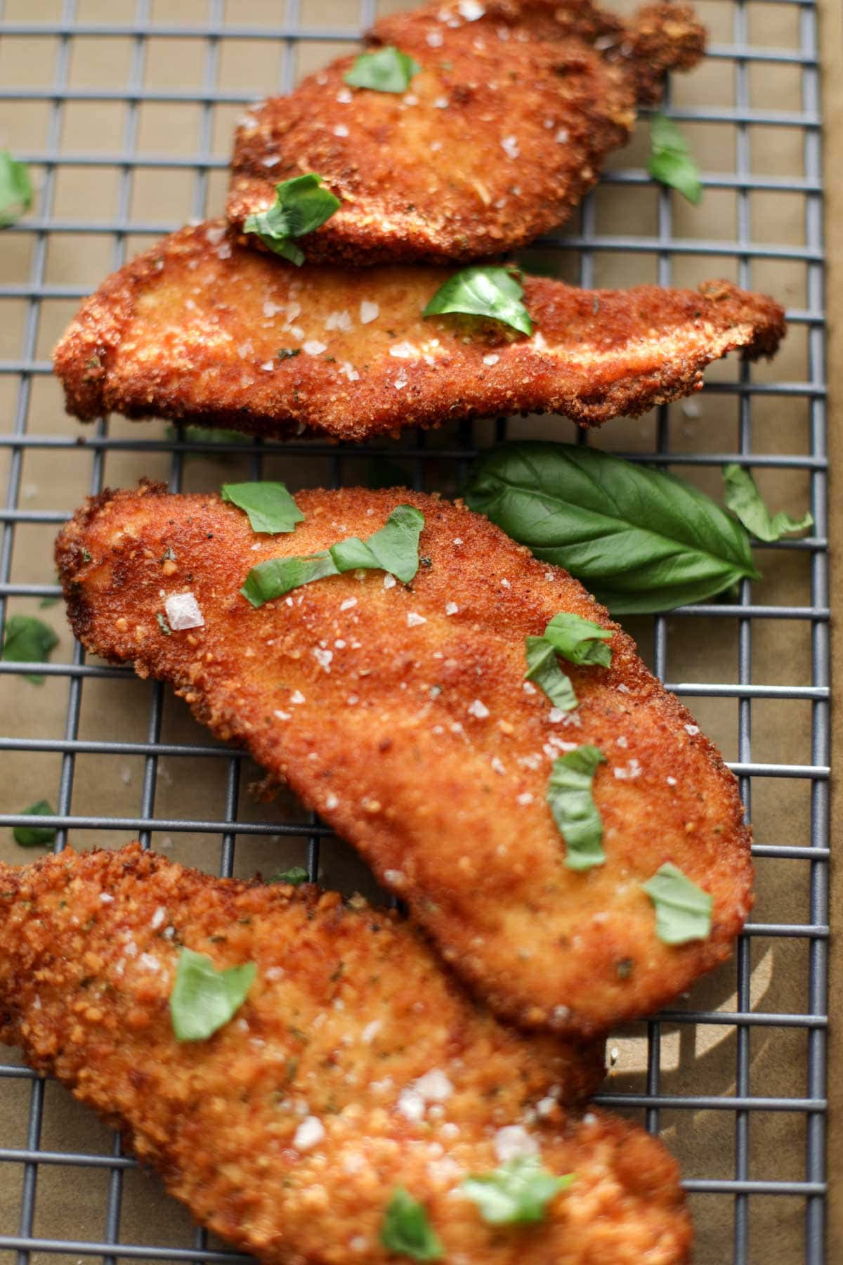 Fried, breaded chicken cutlets on a wire rack topped with chopped basil and malden salt. 