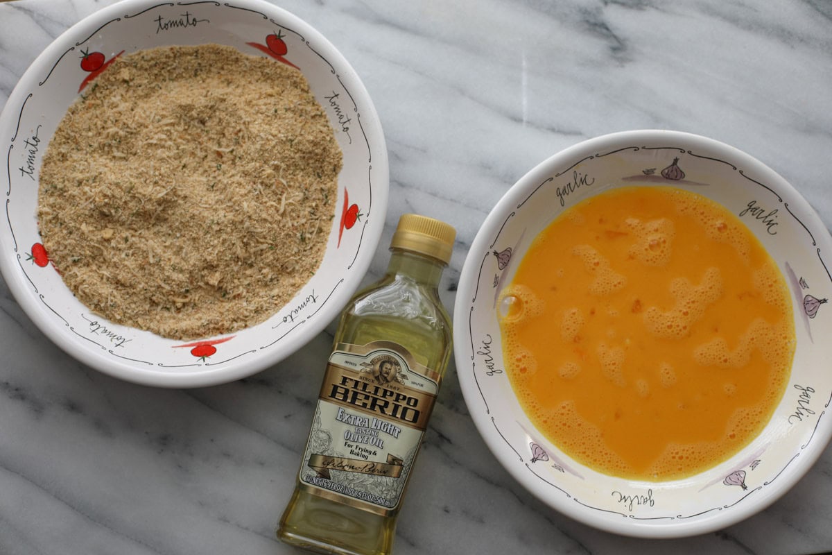 A bowl of seasoned Italian breadcrumbs, a bottle of light extra virgin olive oil, and a bowl of scrambled eggs sitting on a marble counter.