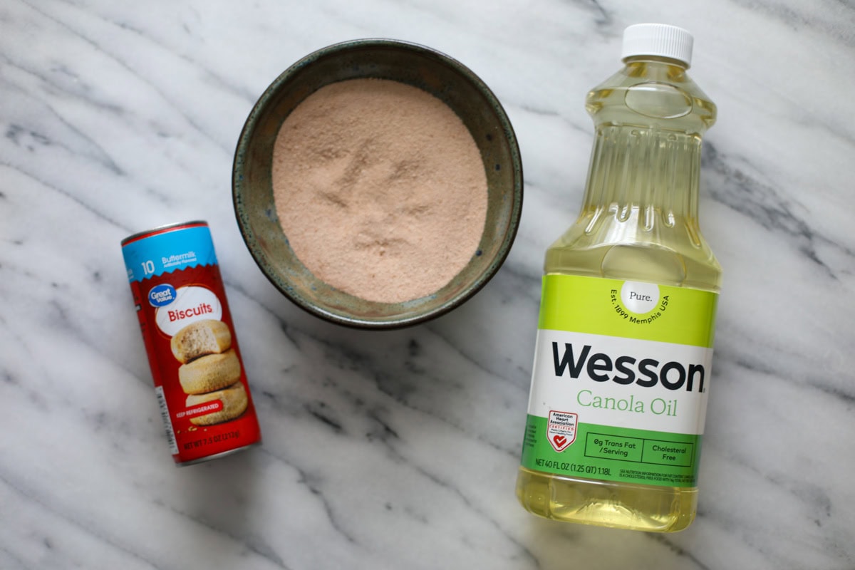 A blue bowl filled with cinnamon and sugar, a bottle of canola oil, and can of ready biscuit dough on a marble counter.