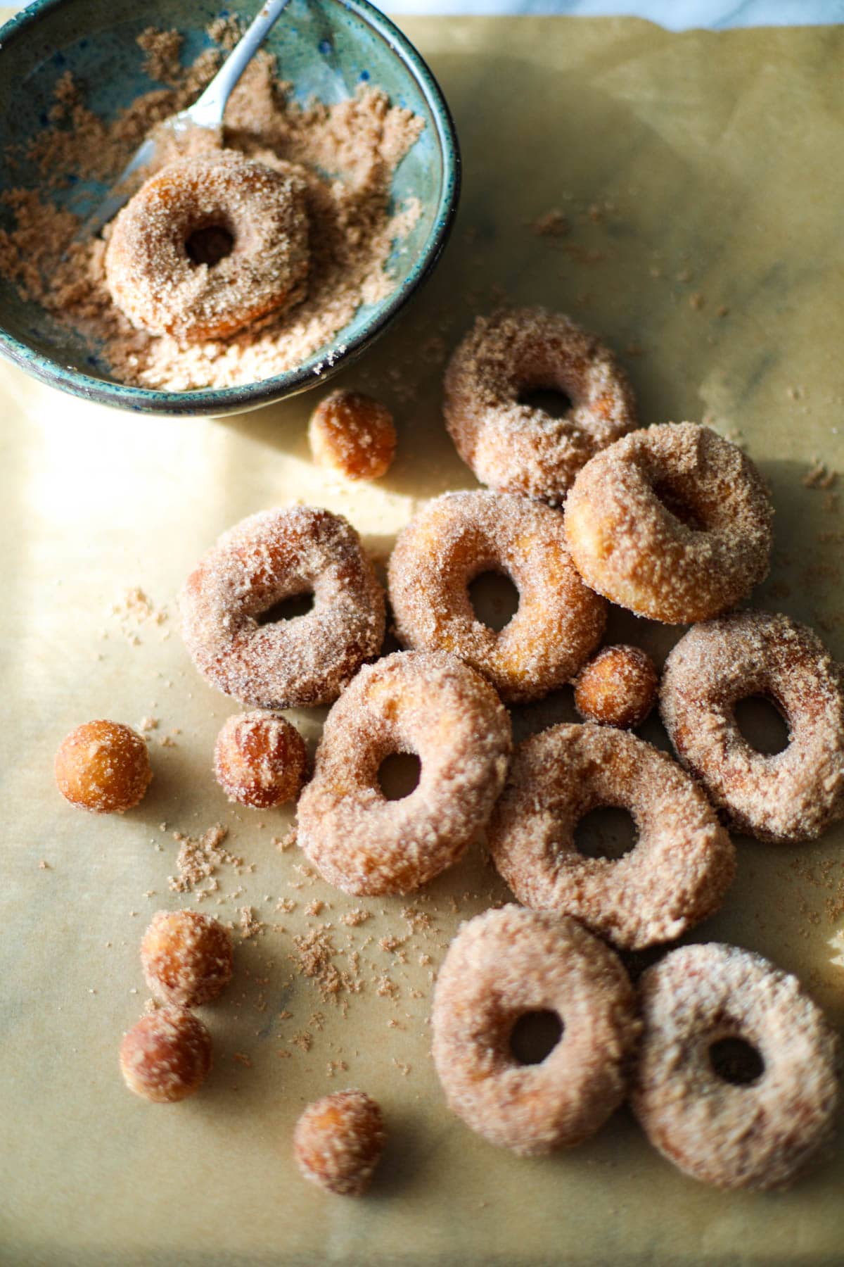 A blue bowl filled with cinnamon and sugar and 10 mini donuts and donut holes on brown parchment paper. 