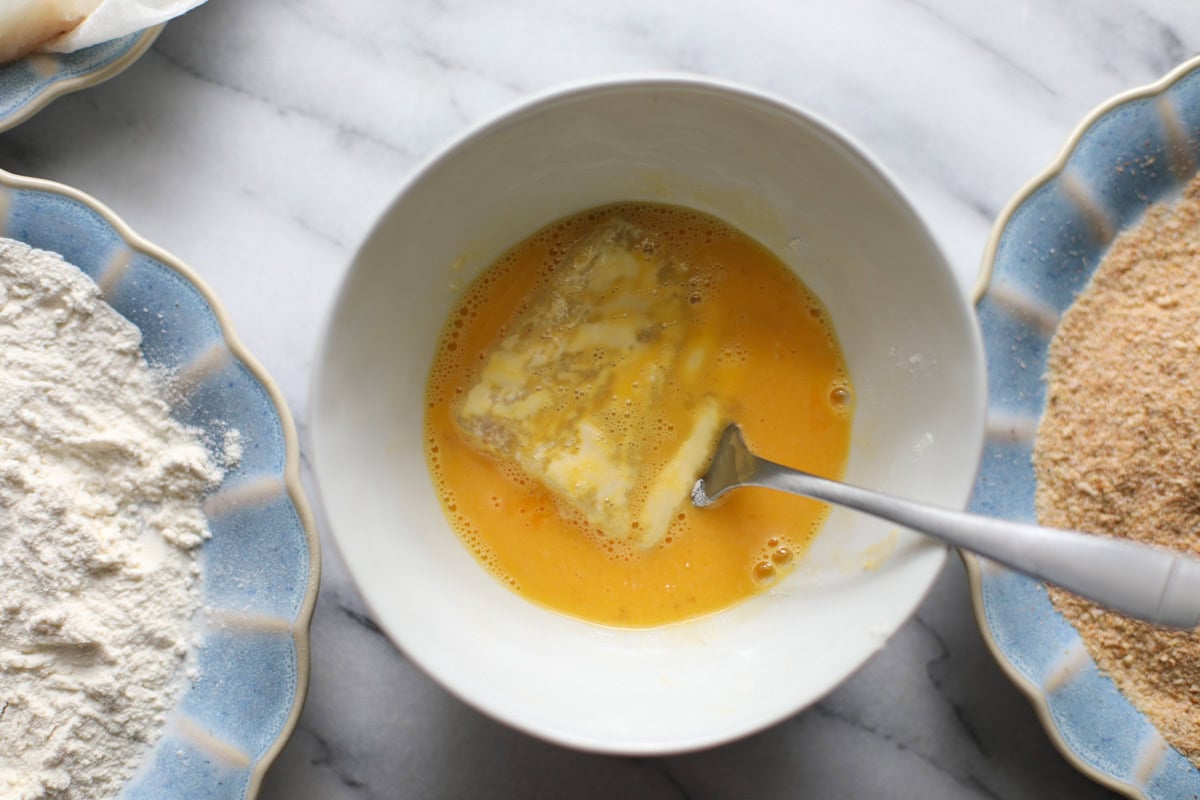 A small white bowl filled with a scrambled egg with a cod filet being dipped before going into breadcrumbs to make a fried fish sandwich.