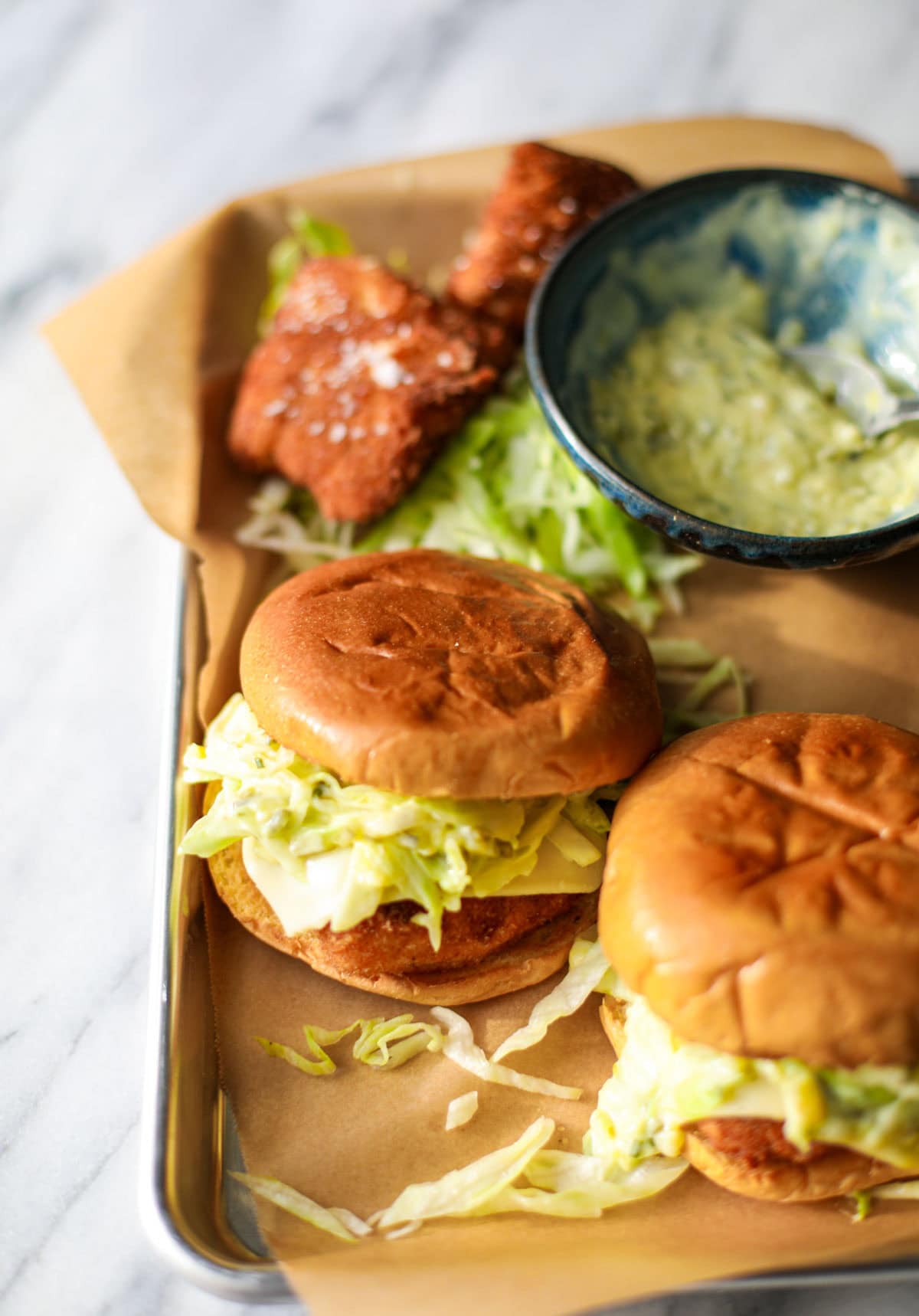 A silver sheet tray with two brioche bun fish sandwiches topped with lettuce and cheese with a blue bowl of tartar sauce on the side with a metal spoon.