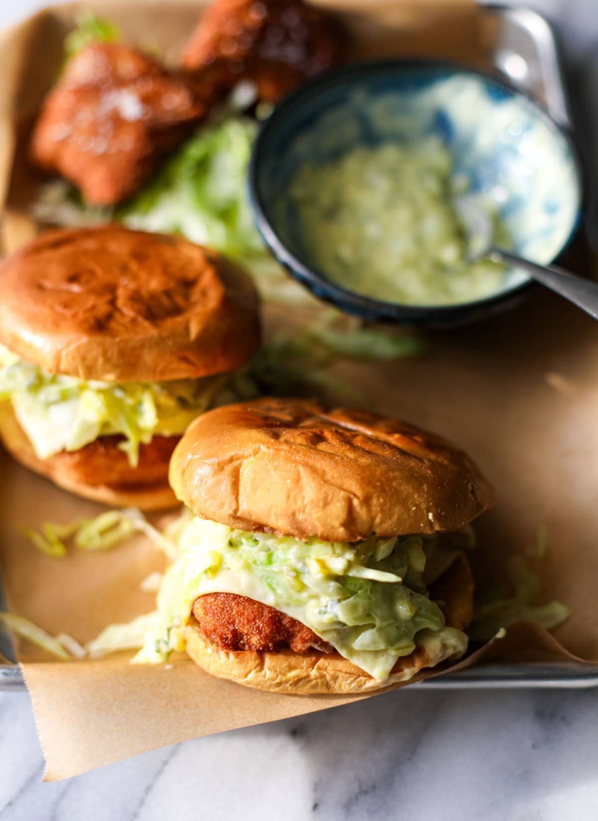 A silver sheet tray with two brioche bun fried fish sandwiches topped with lettuce and cheese with a  blue bowl of tartar sauce on the side with a metal spoon.